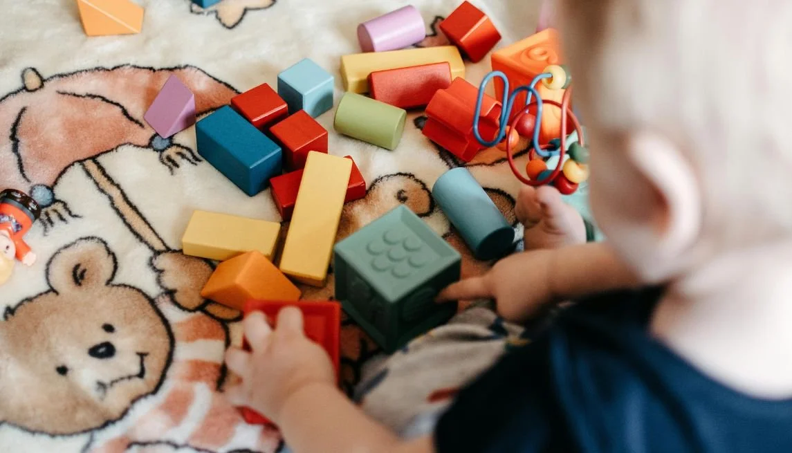 Child playing with colorful building blocks and toys on a cartoon bear-themed rug.