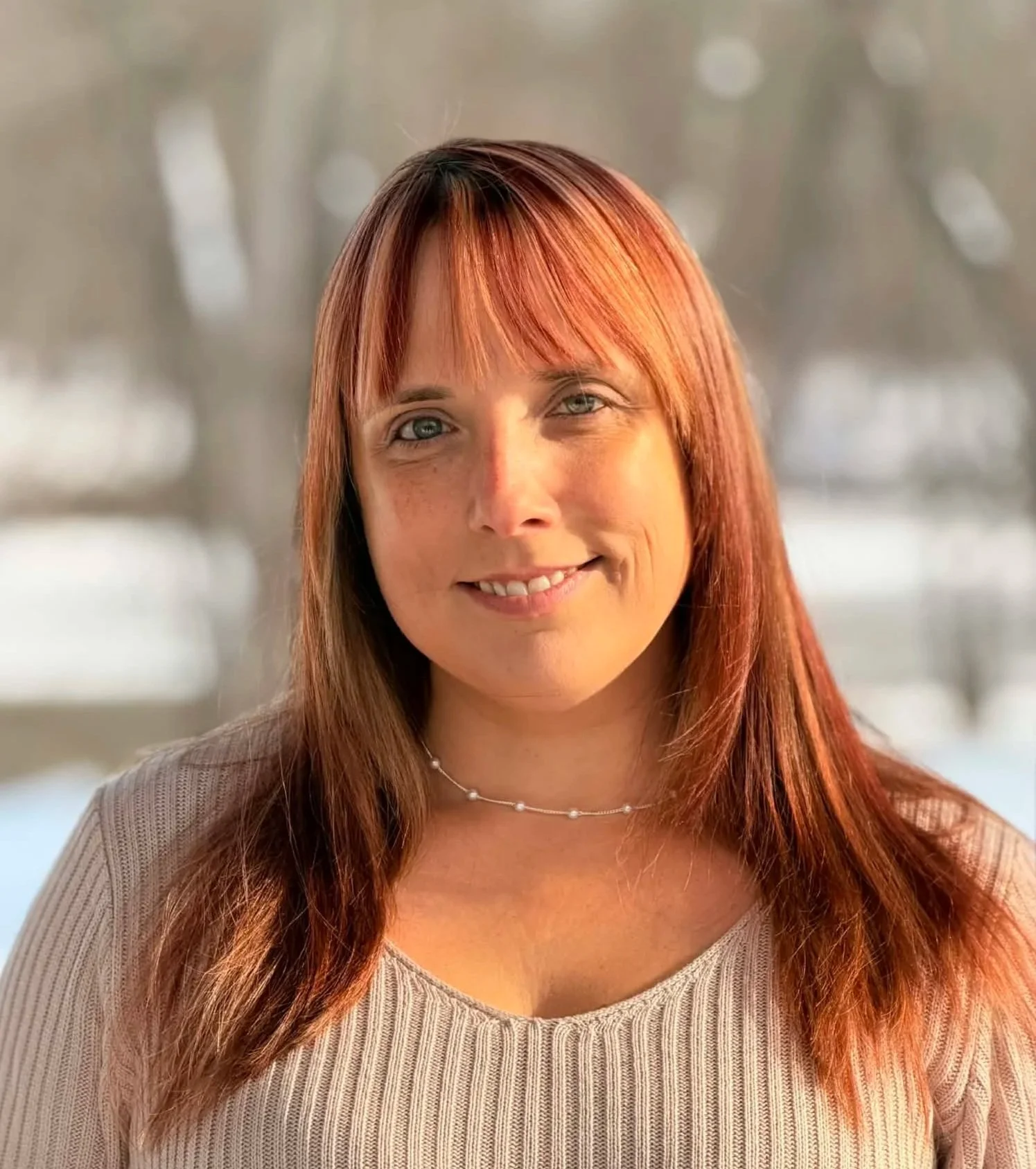 Amber Johnson, owner of Miss Amber's Daycare, smiling and outdoors in winter, wearing a beige sweater and a pearl choker necklace, with snow and trees in the background.