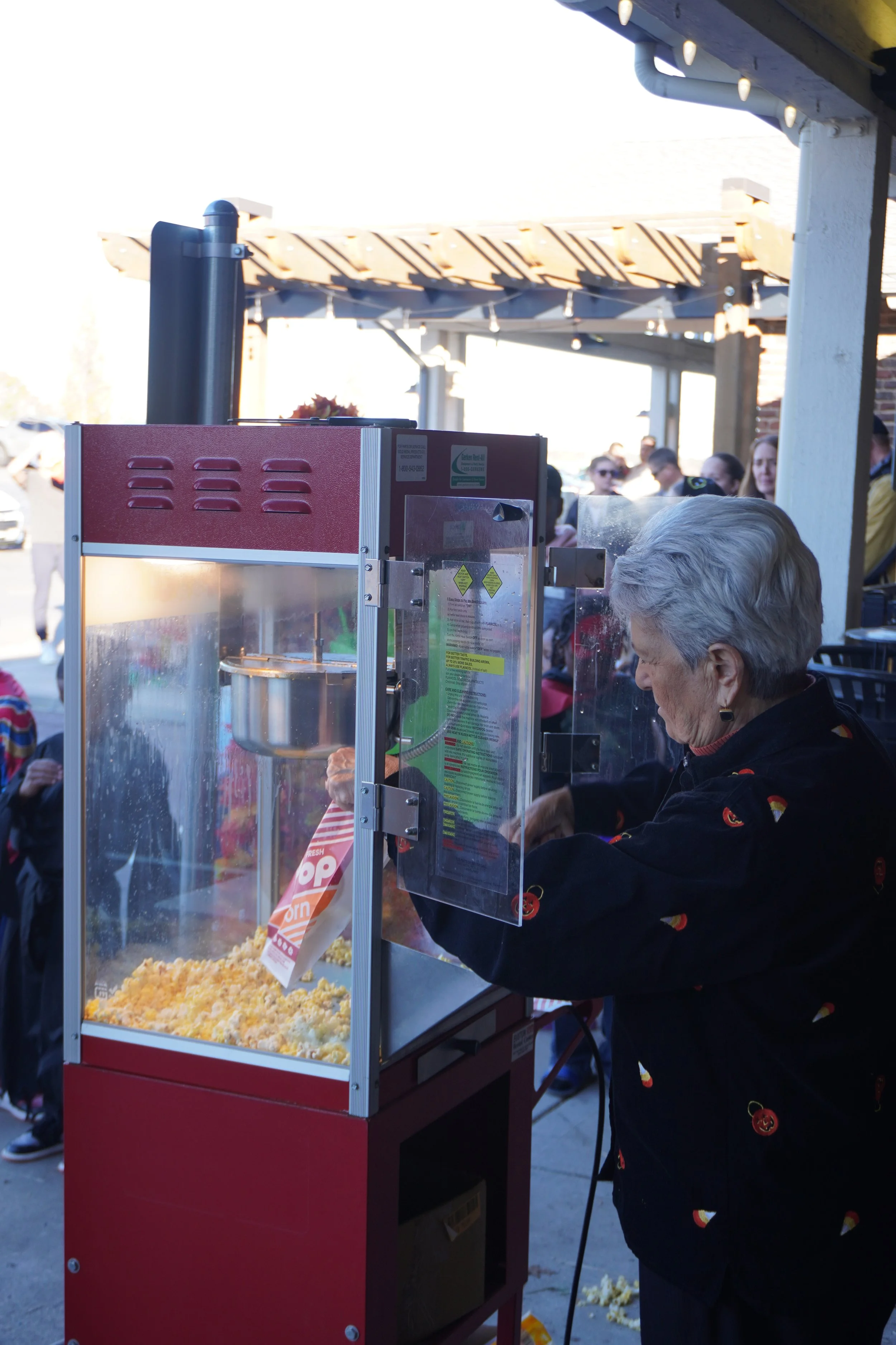 An elderly woman operating a red popcorn machine outdoors with a crowd of people in the background.