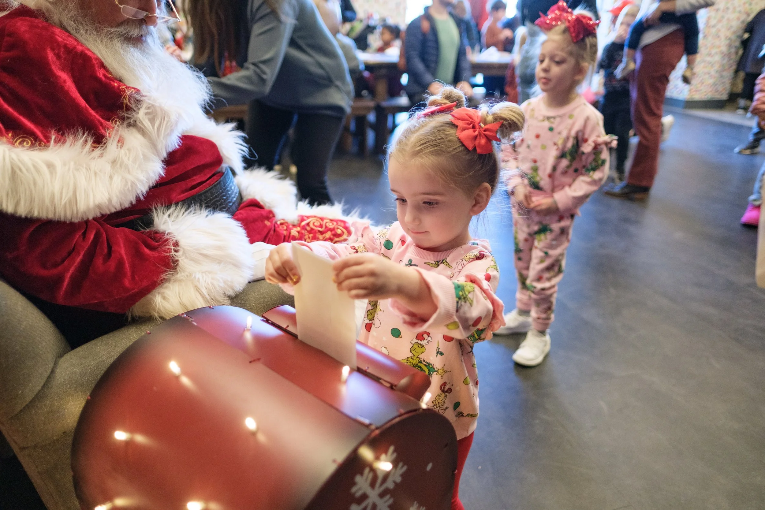 A young girl in a Christmas-themed pink sweater and red pants is opening a present from Santa Claus, who is dressed in a traditional red and white suit. Other children and adults are in the background, some dressed in pajamas, in a festive indoor set