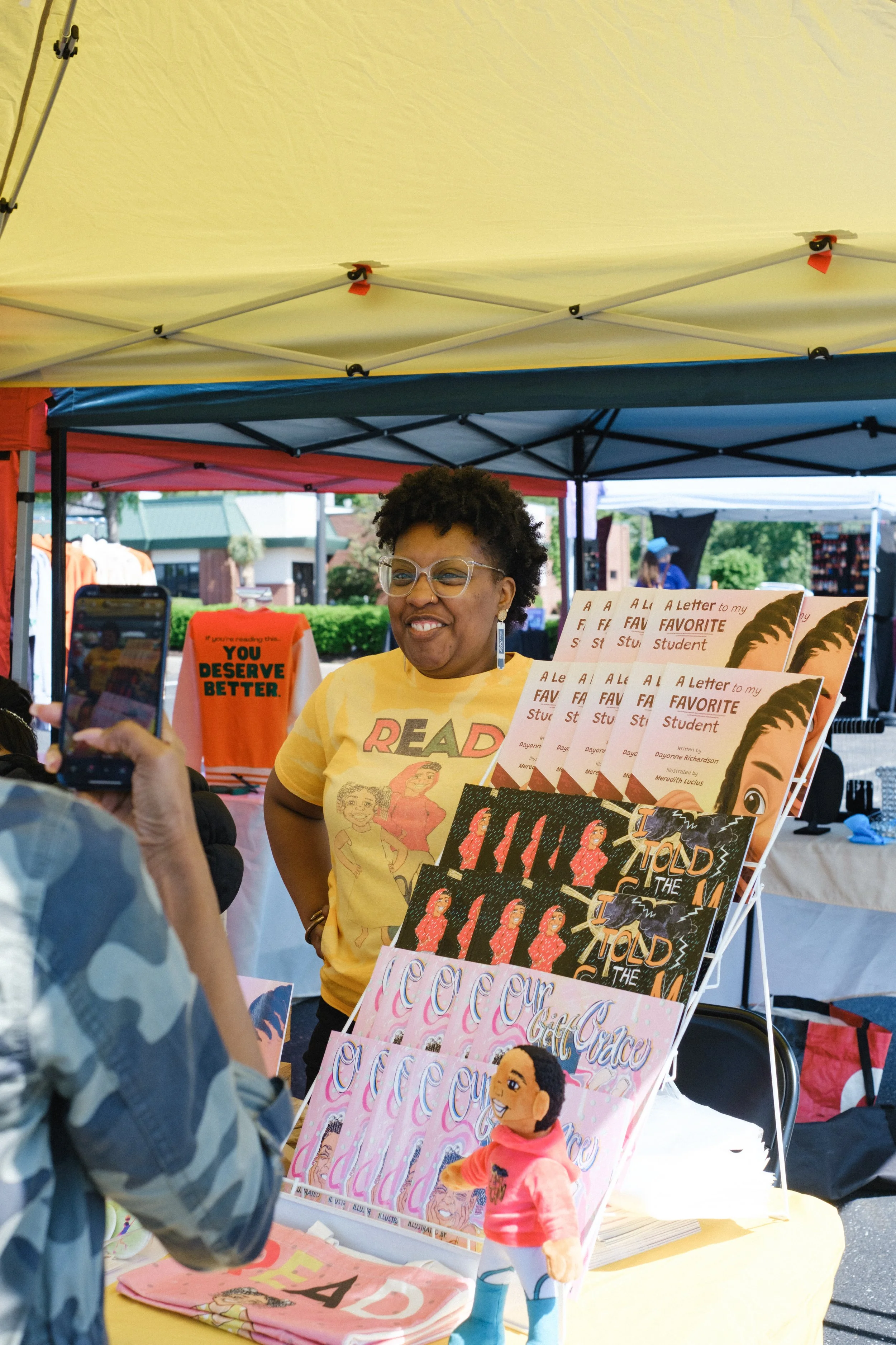 Woman standing behind a table with children's books and a doll at an outdoor event, smiling, with a yellow canopy tent overhead.