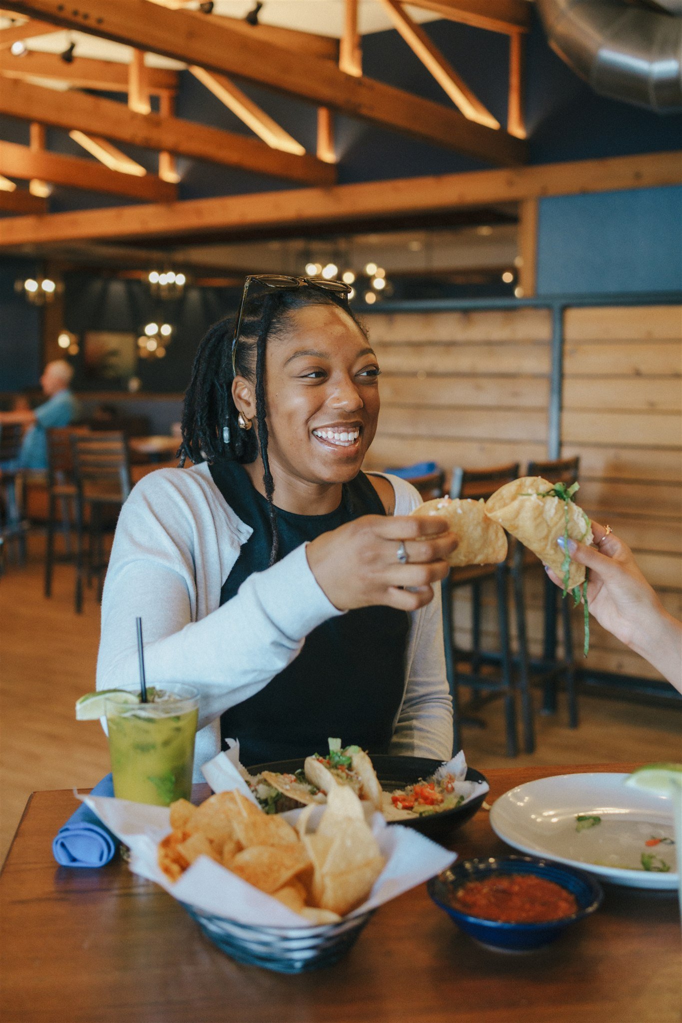 Smiling woman with glasses on her head and dreadlocks enjoying a taco at a restaurant with wood accents and dim lighting, with chips, salsa, and a drink on the table.