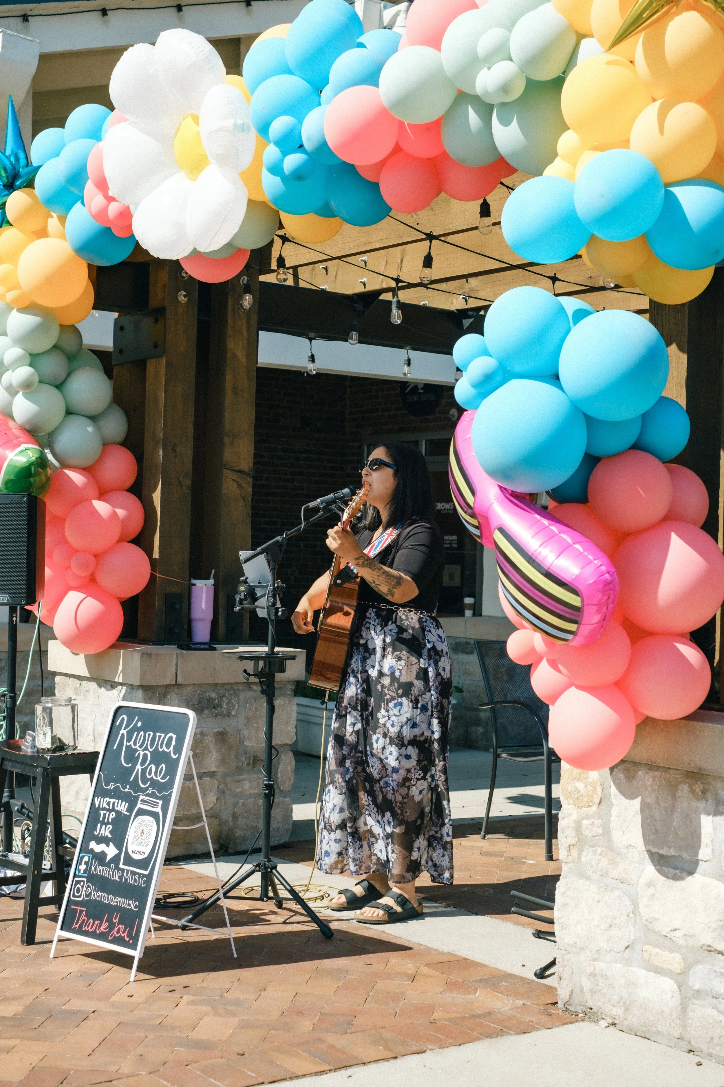 A woman performing with a guitar under a balloon arch at an outdoor event, with a signboard displaying her name and social media handles.