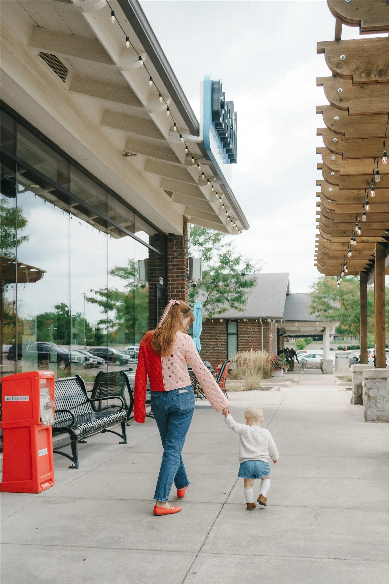 A woman holding the hand of a young child while walking outside an outdoor shopping plaza with storefronts, trees, and parked cars in the background.