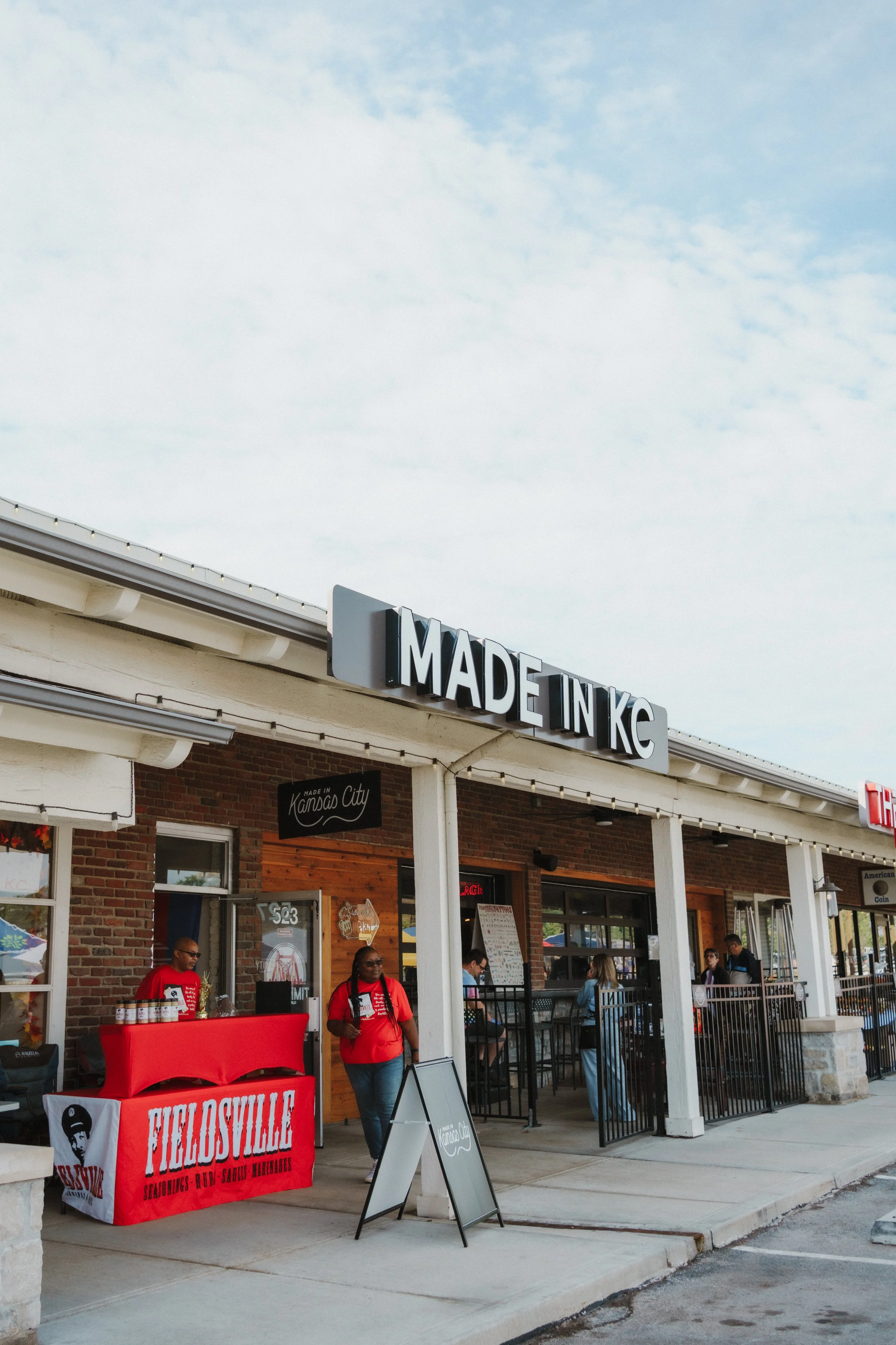 Exterior view of a shopping or dining area with a sign that says 'Made in KC' on a brick building. There are people standing and sitting outside, some wearing red shirts and a table with a red cloth that has the words 'Fidelville' on it.