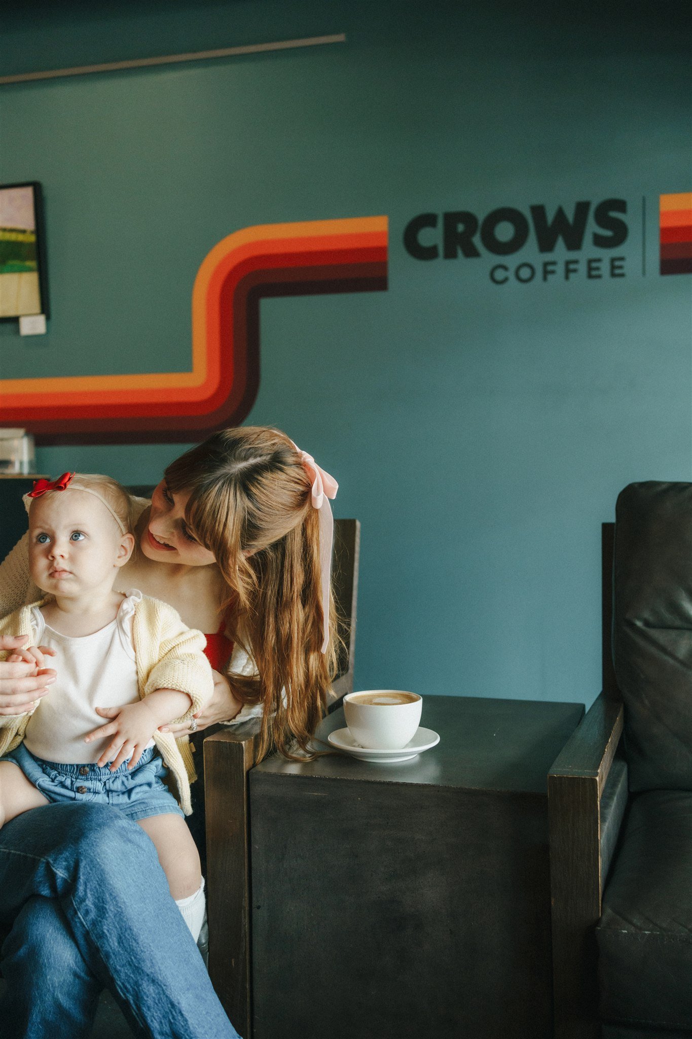 A woman with long red hair and a pink bow in her hair is sitting with a young girl in a coffee shop called Crows Coffee. The girl is wearing a white shirt, blue shorts, and a yellow cardigan. A coffee cup with a latte is on the table beside them. The