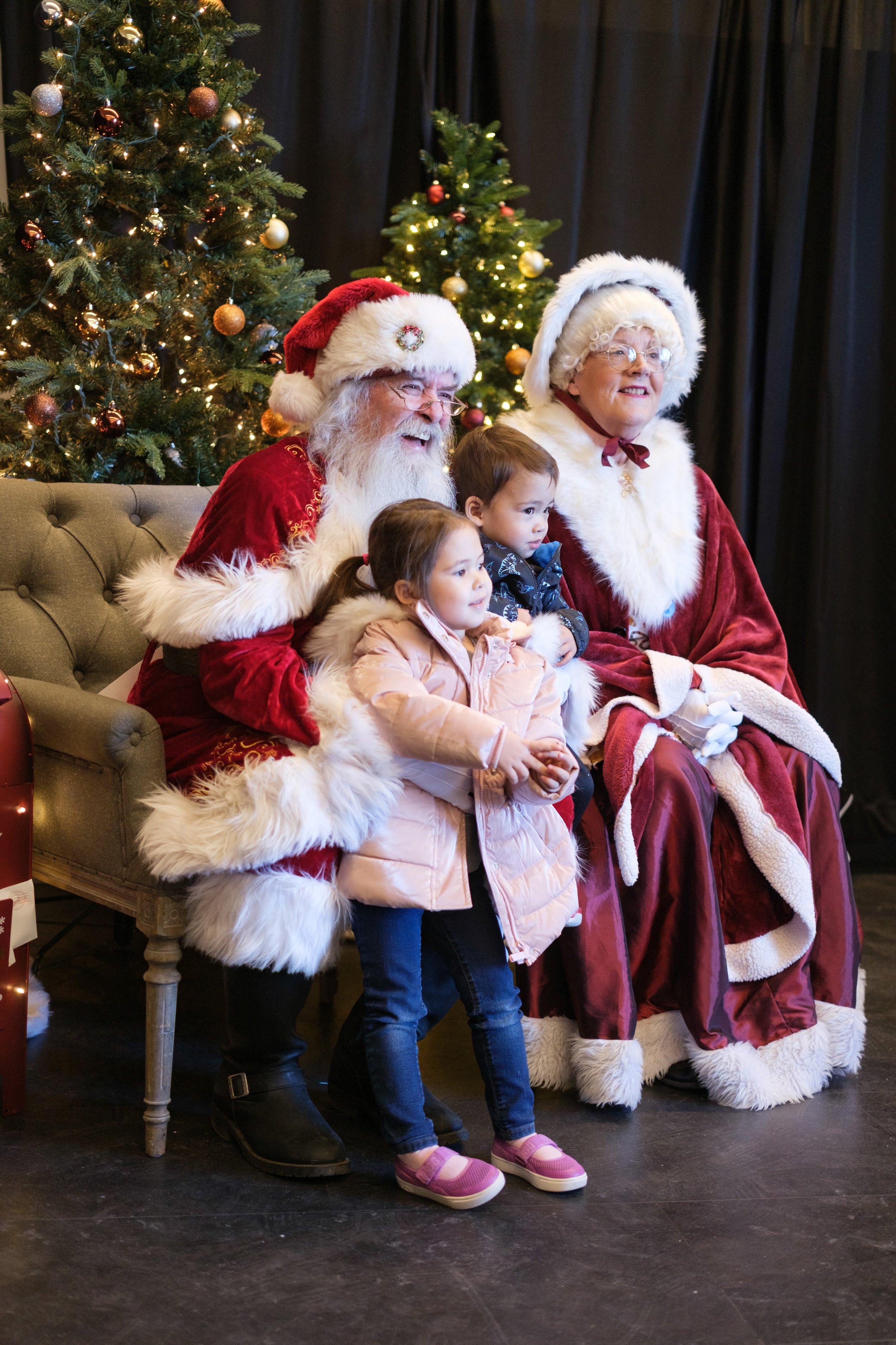 Children sitting with Santa Claus and Mrs. Claus during a Christmas event, with decorated Christmas trees in the background.