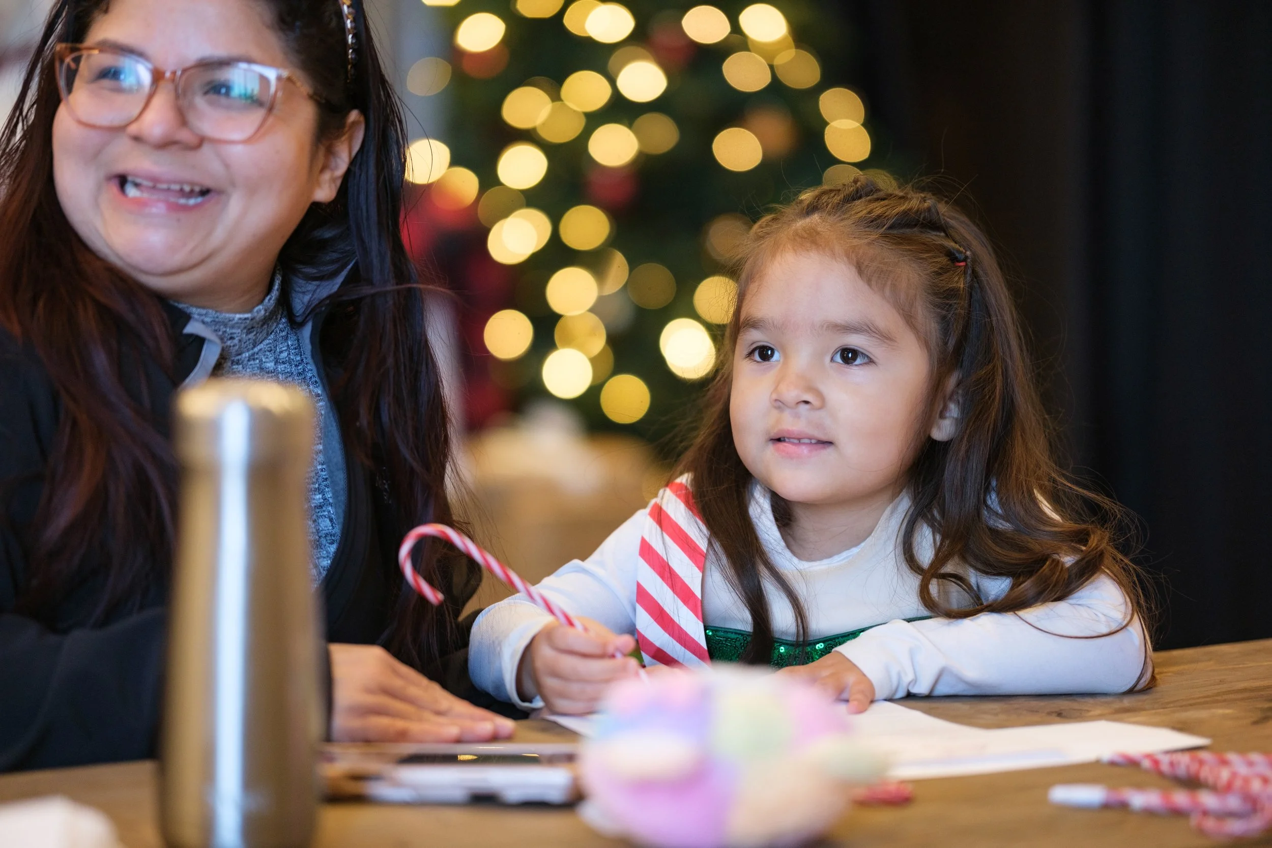 A woman and a young girl sitting at a table with Christmas decorations in the background, smiling and enjoying holiday activities.