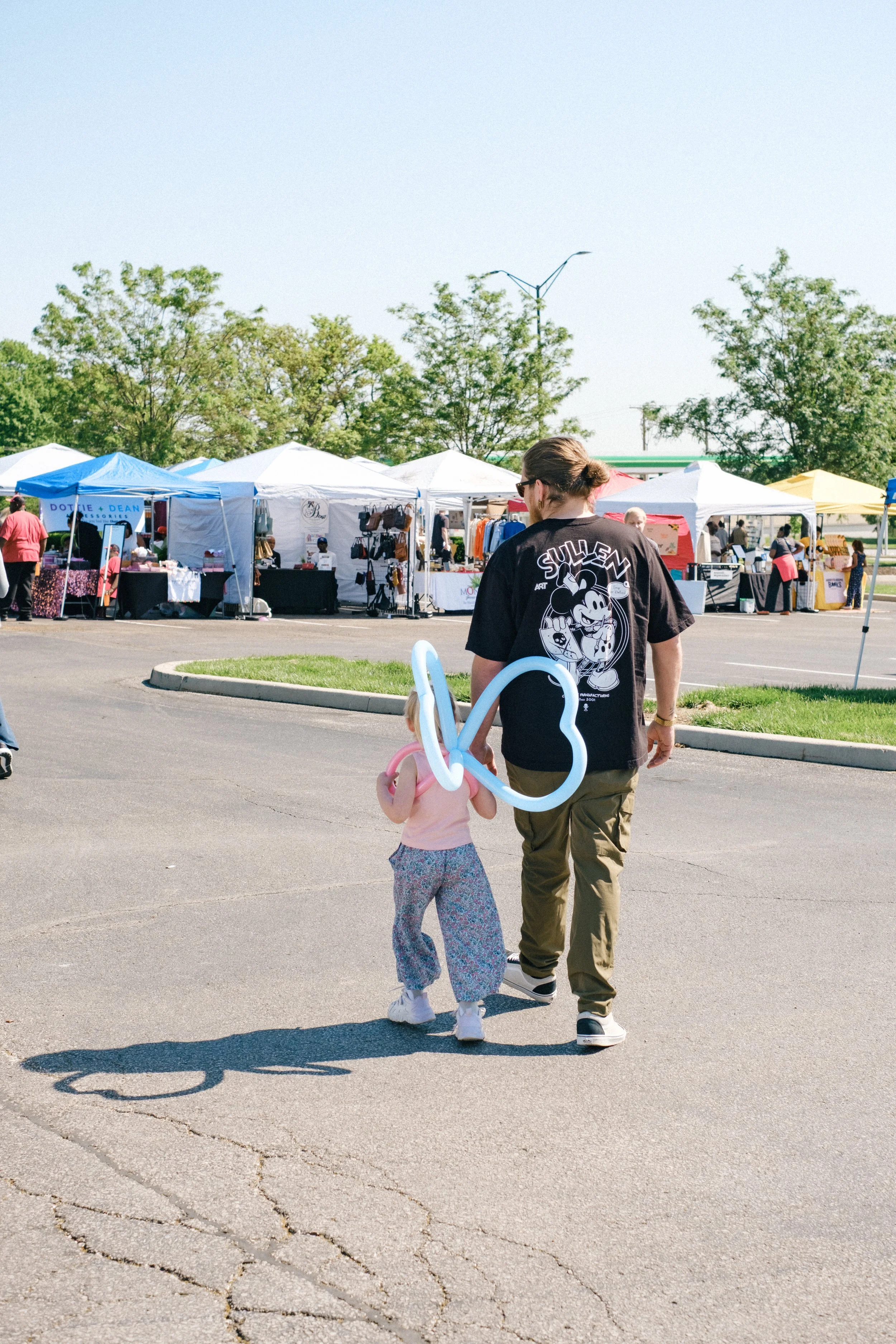 A man and a young girl walking through an outdoor market, with market stalls and trees in the background. The girl is holding balloon animals.