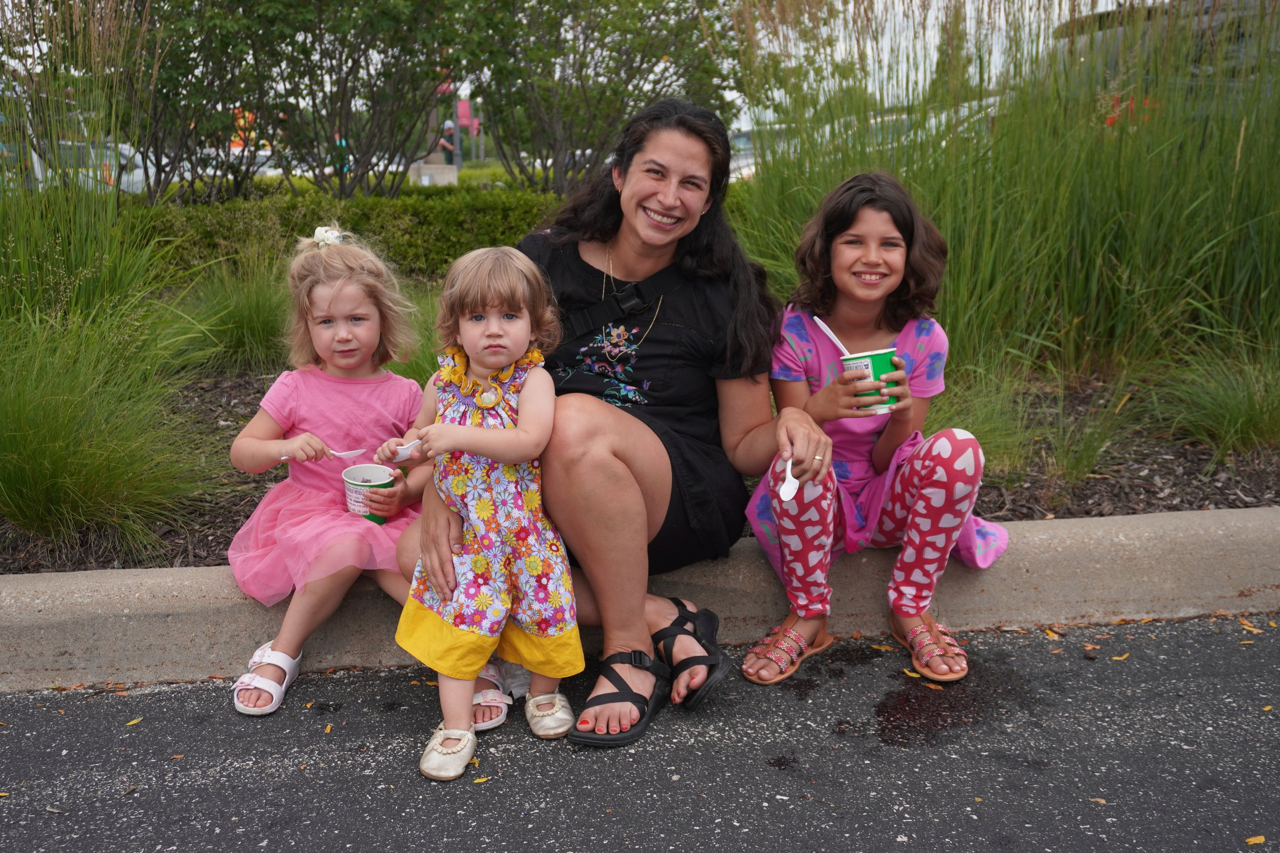 Four young girls and a woman sitting on a curb in a park, eating cups of frozen yogurt, with greenery and trees in the background.