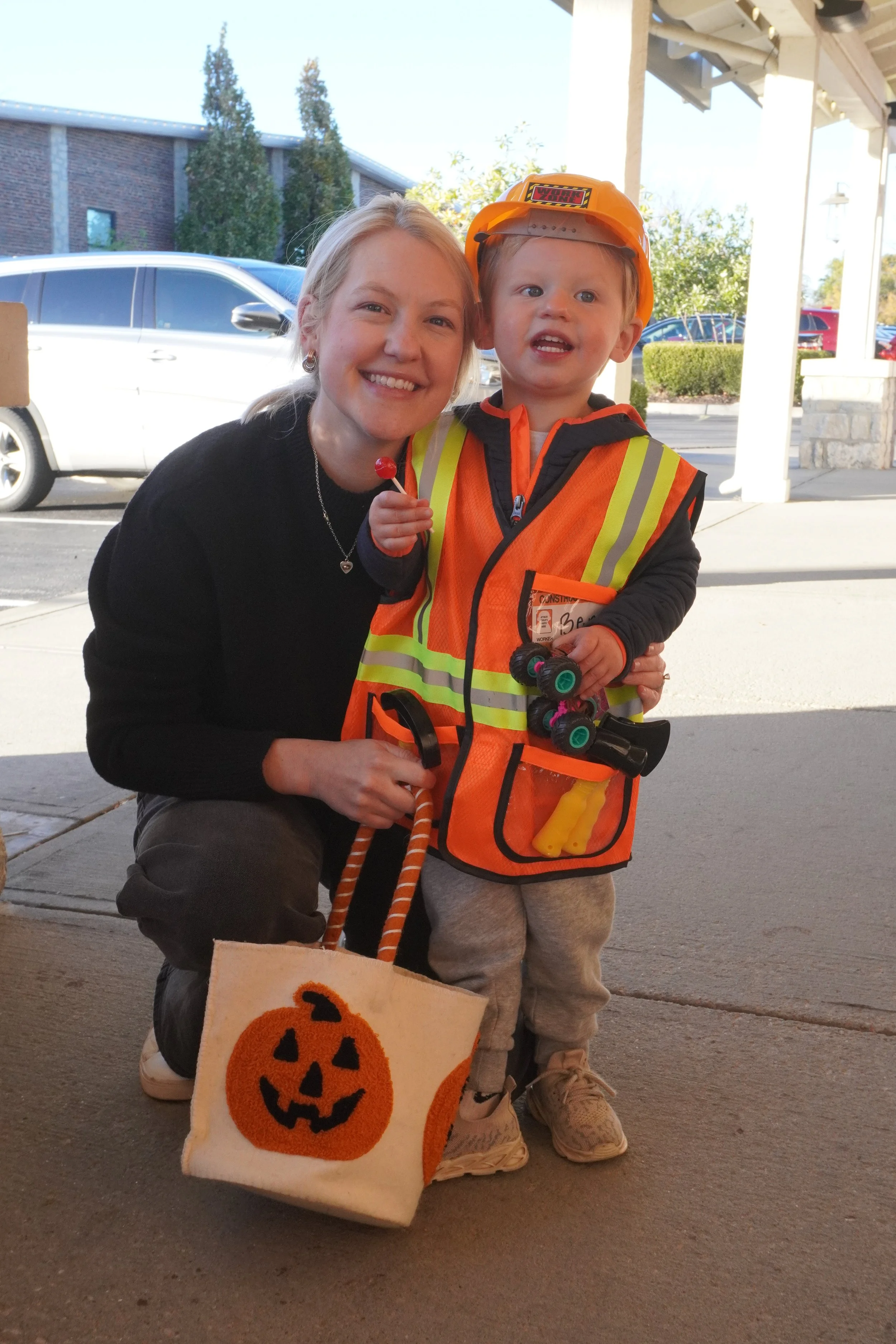 A smiling woman and a young child dressed in construction gear, holding a Halloween-themed bag with a pumpkin face, outdoors in front of parked cars and a building.
