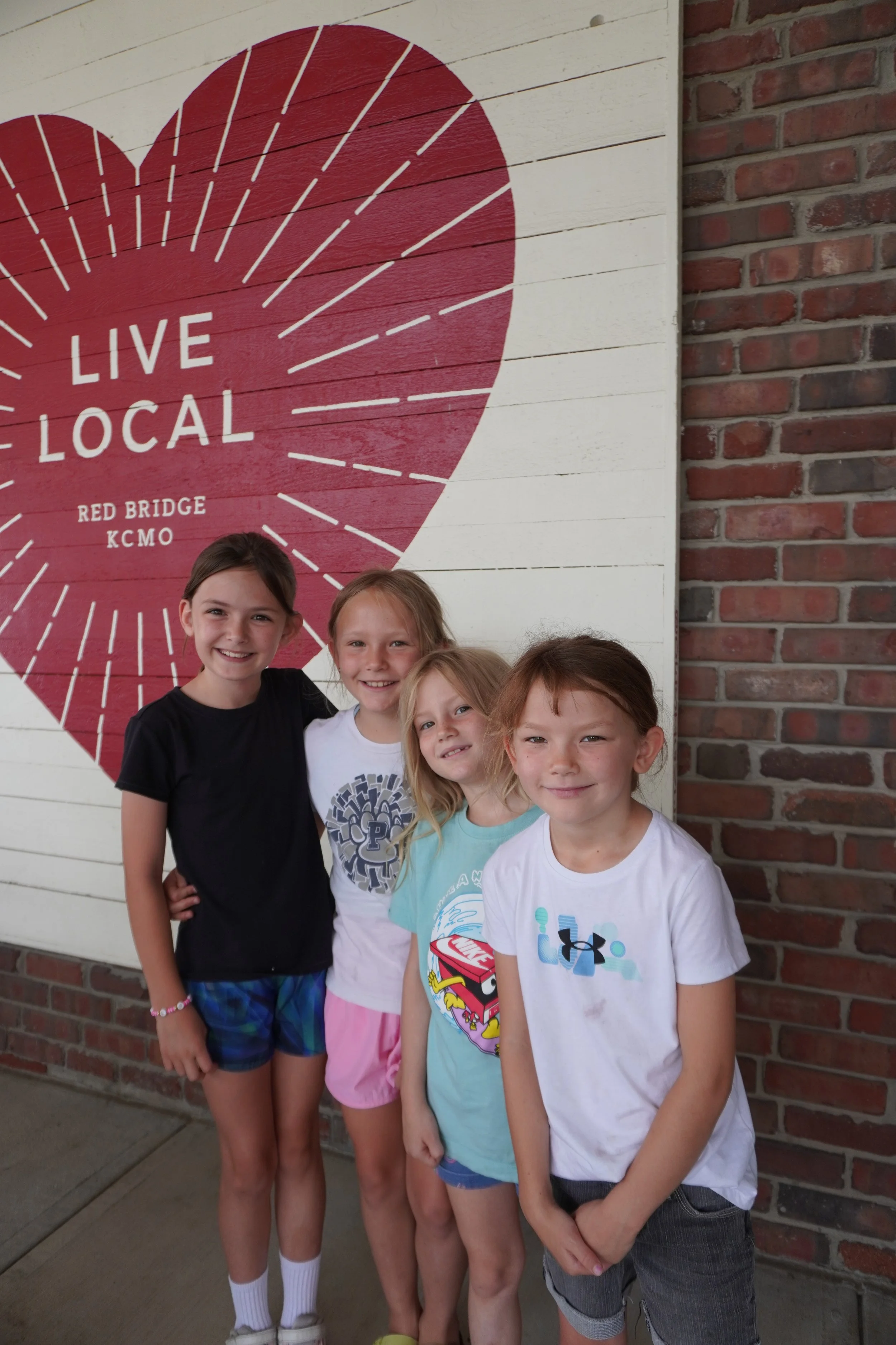 Four young girls standing in front of a painted sign that says "Live Local" with a heart design and the location names "Red Bridge" and "KCMO." The girls are smiling, dressed casually, and appear to be enjoying a day out.