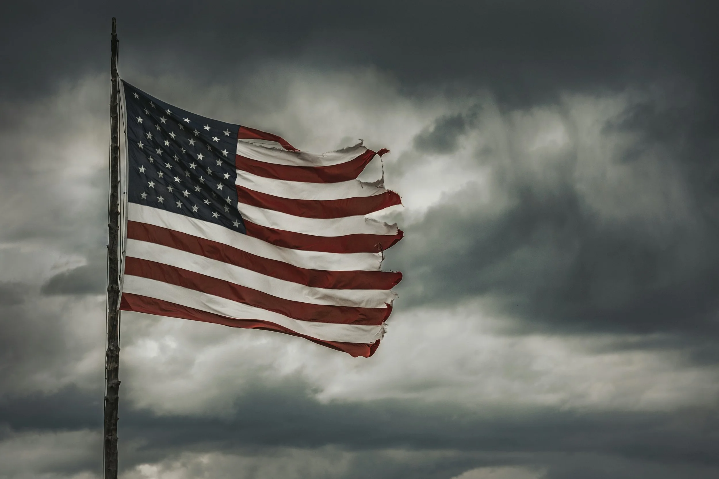 A worn American flag blowing against a dark, cloudy sky.