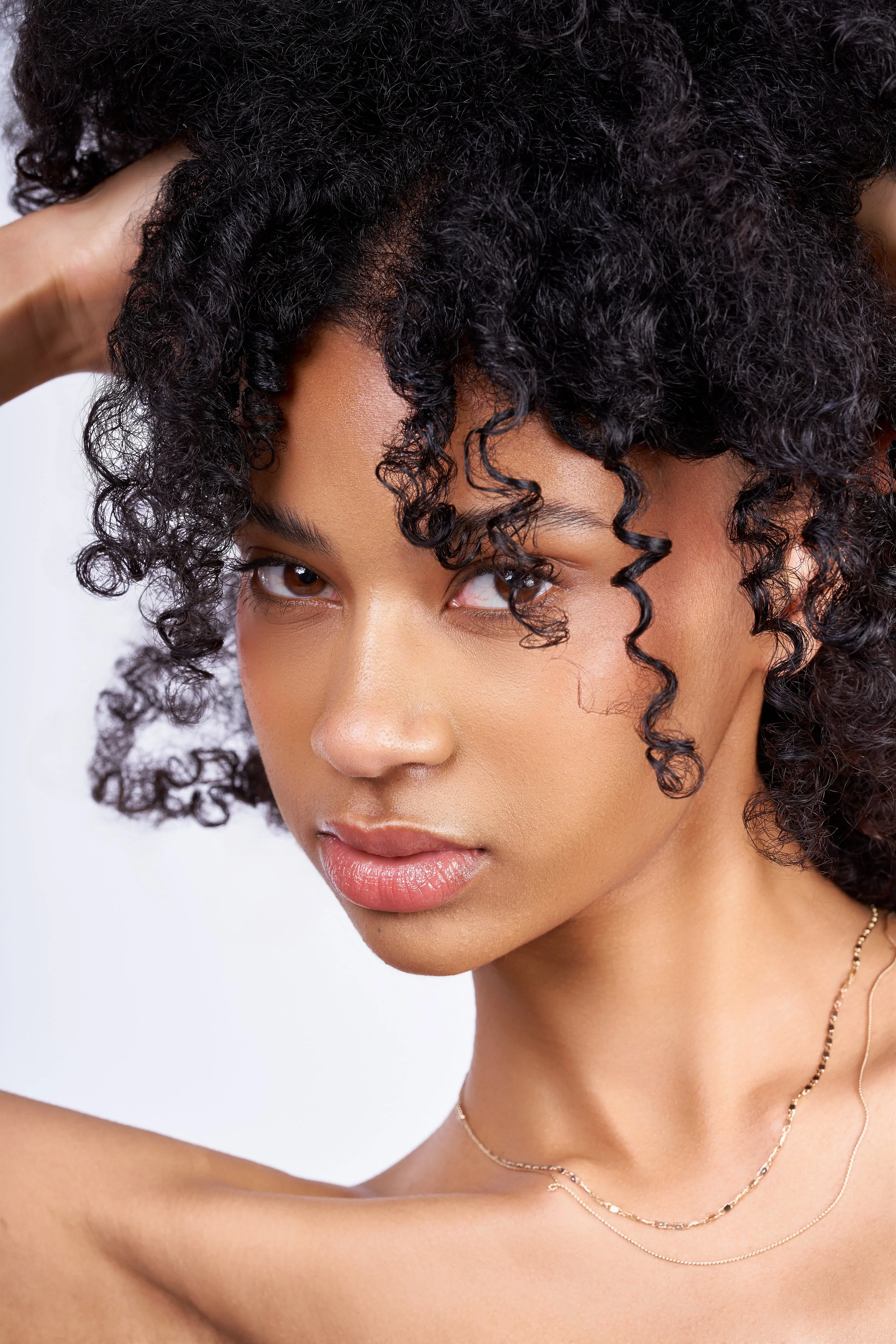 A young woman with dark, curly hair and smooth skin, wearing delicate gold necklaces, posing with her hand behind her head against a plain white background.