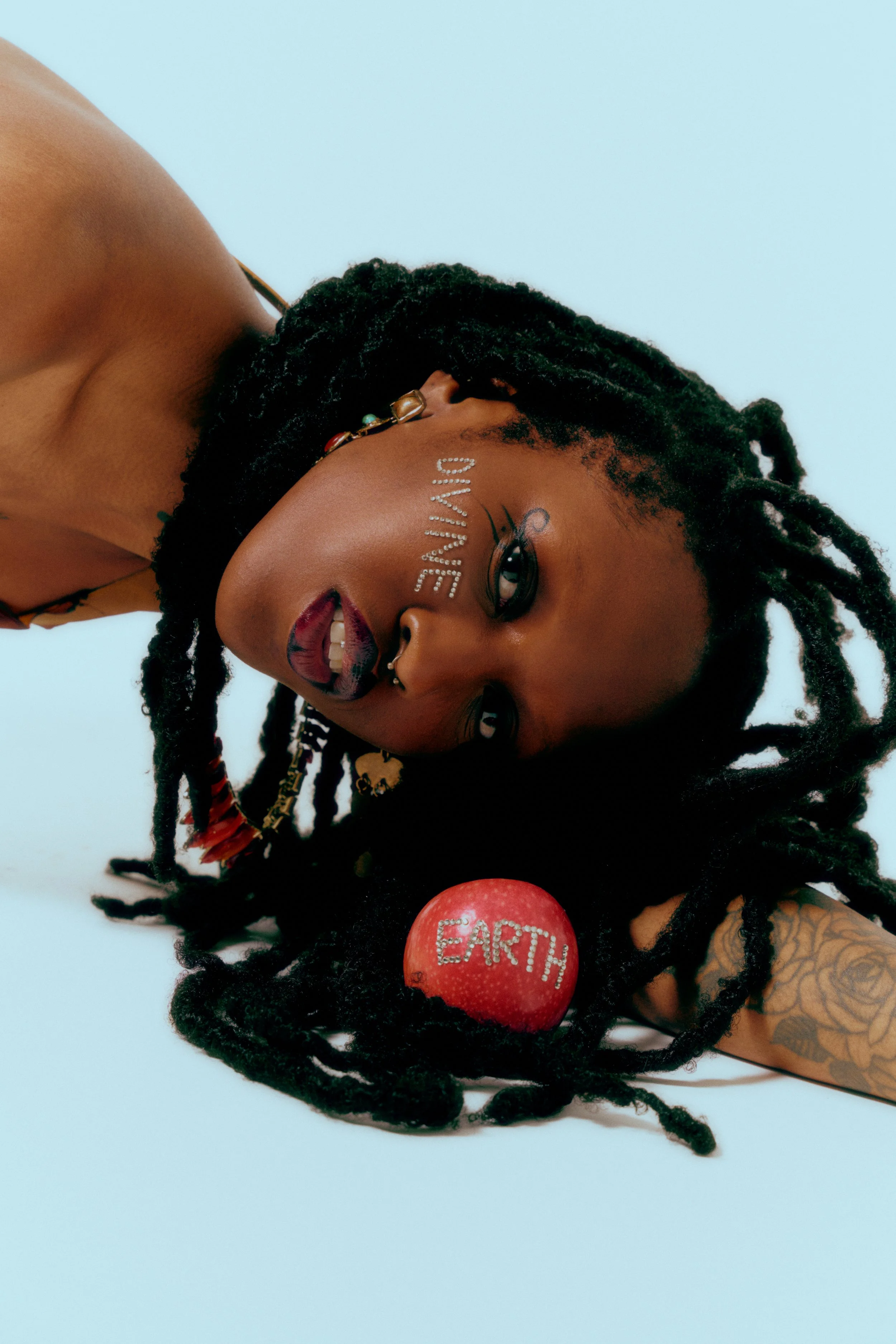 A person with long dreadlocks lying down on a light blue background. They have the word "DIVERSITY" written in rhinestones on their forehead, and makeup that emphasizes their eyes and lips. They are holding a red apple with the word "EARTH" written in rhinestones.