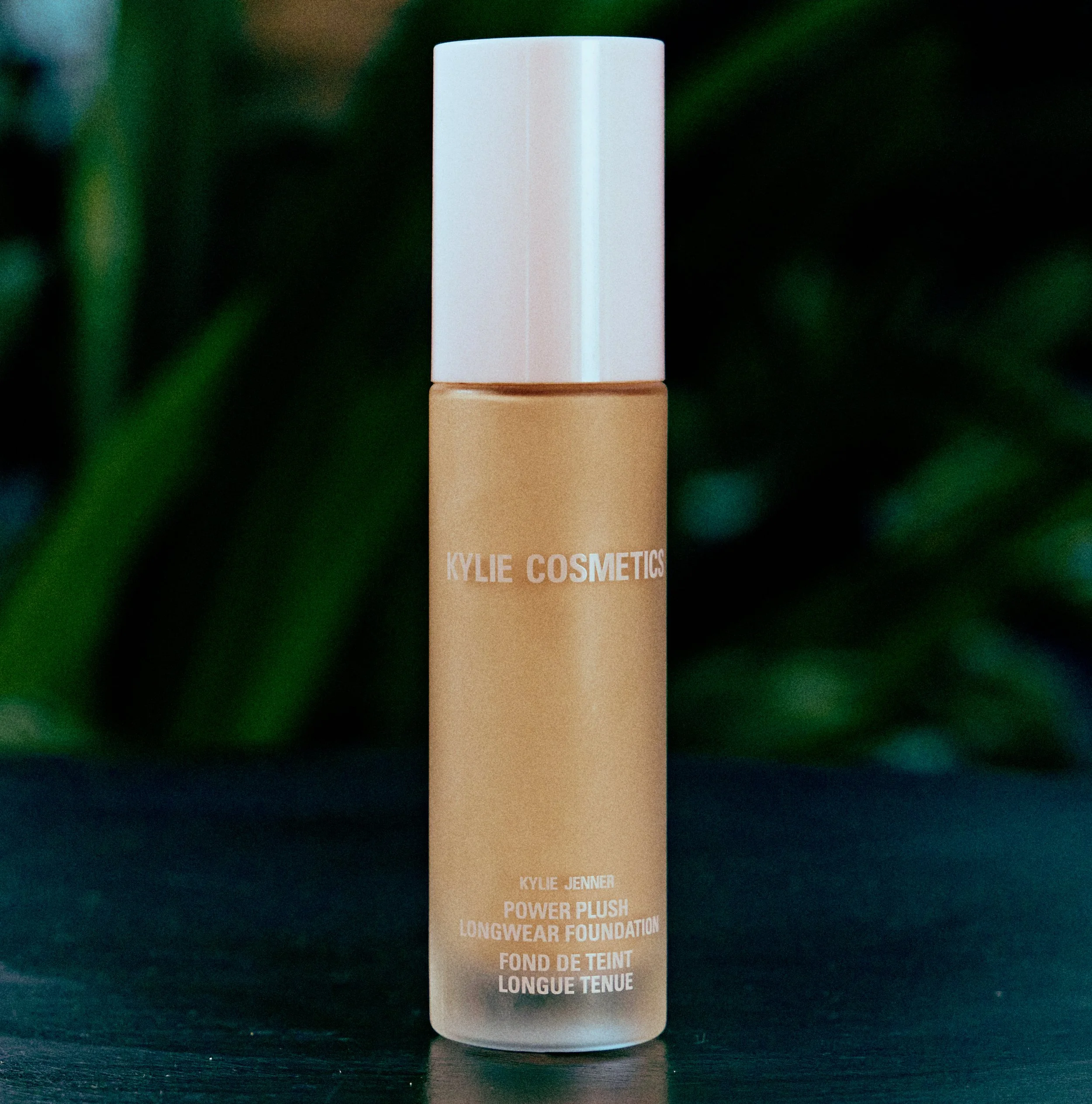Kylie Cosmetics Power Plush Longwear Foundation bottle with a beige cap and a pinkish-beige label, placed on a dark surface with a blurred green background.