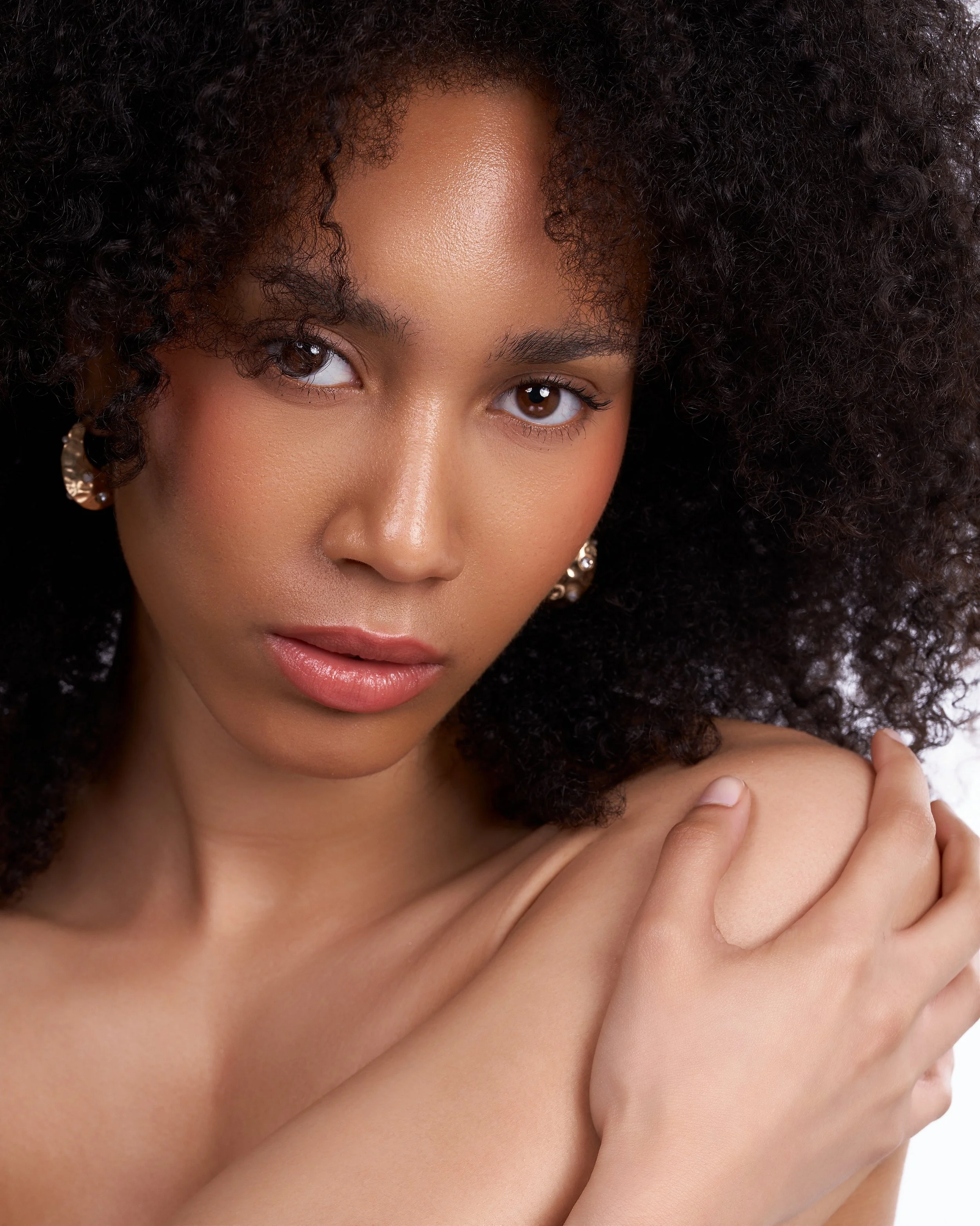 Close-up portrait of a woman with natural curly hair and brown eyes, gently holding her shoulder with her hand, against a white background.