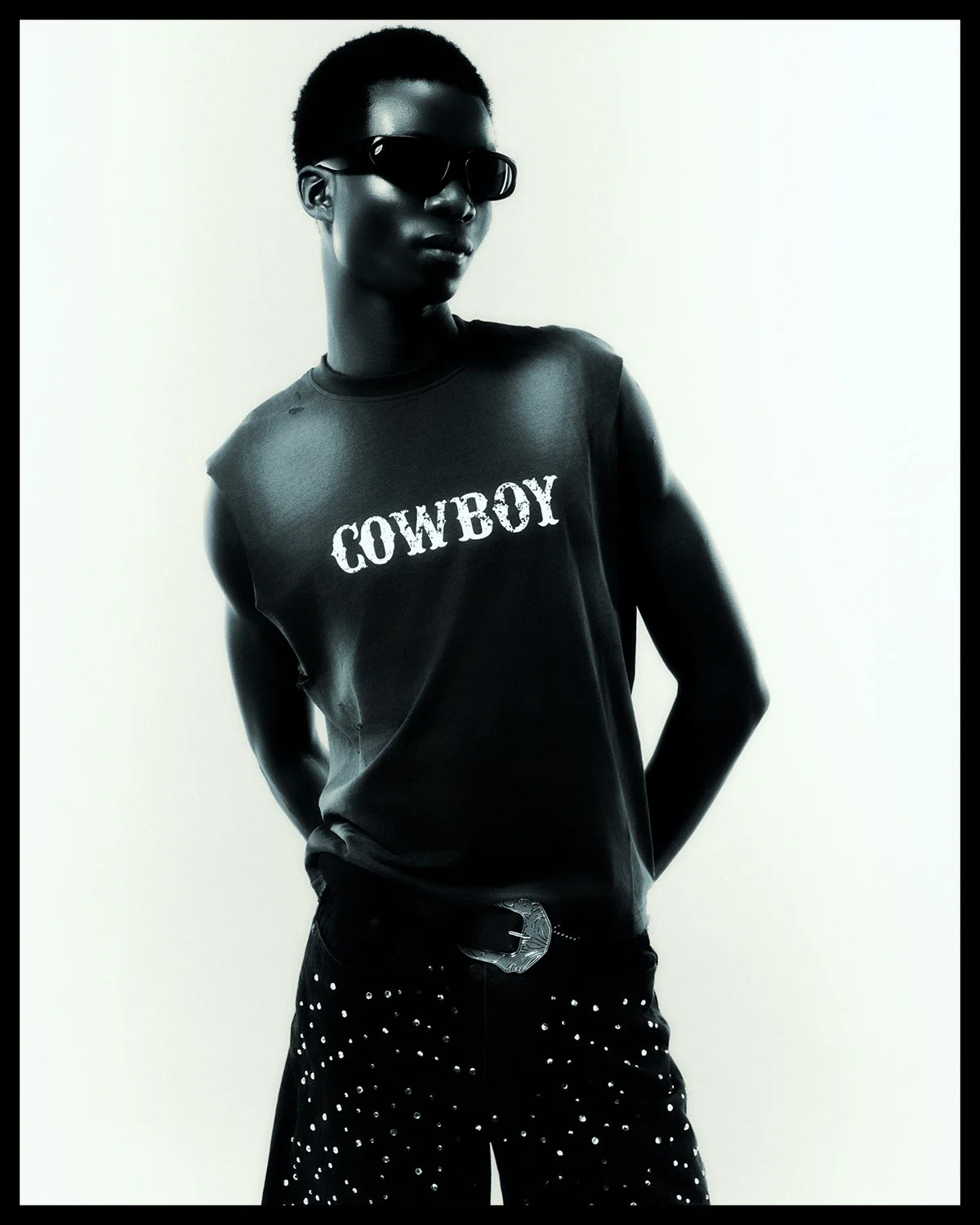 A woman wearing dark sunglasses, a sleeveless black shirt with 'COWBOY' written on it, and black pants with white dots, standing against a plain background.