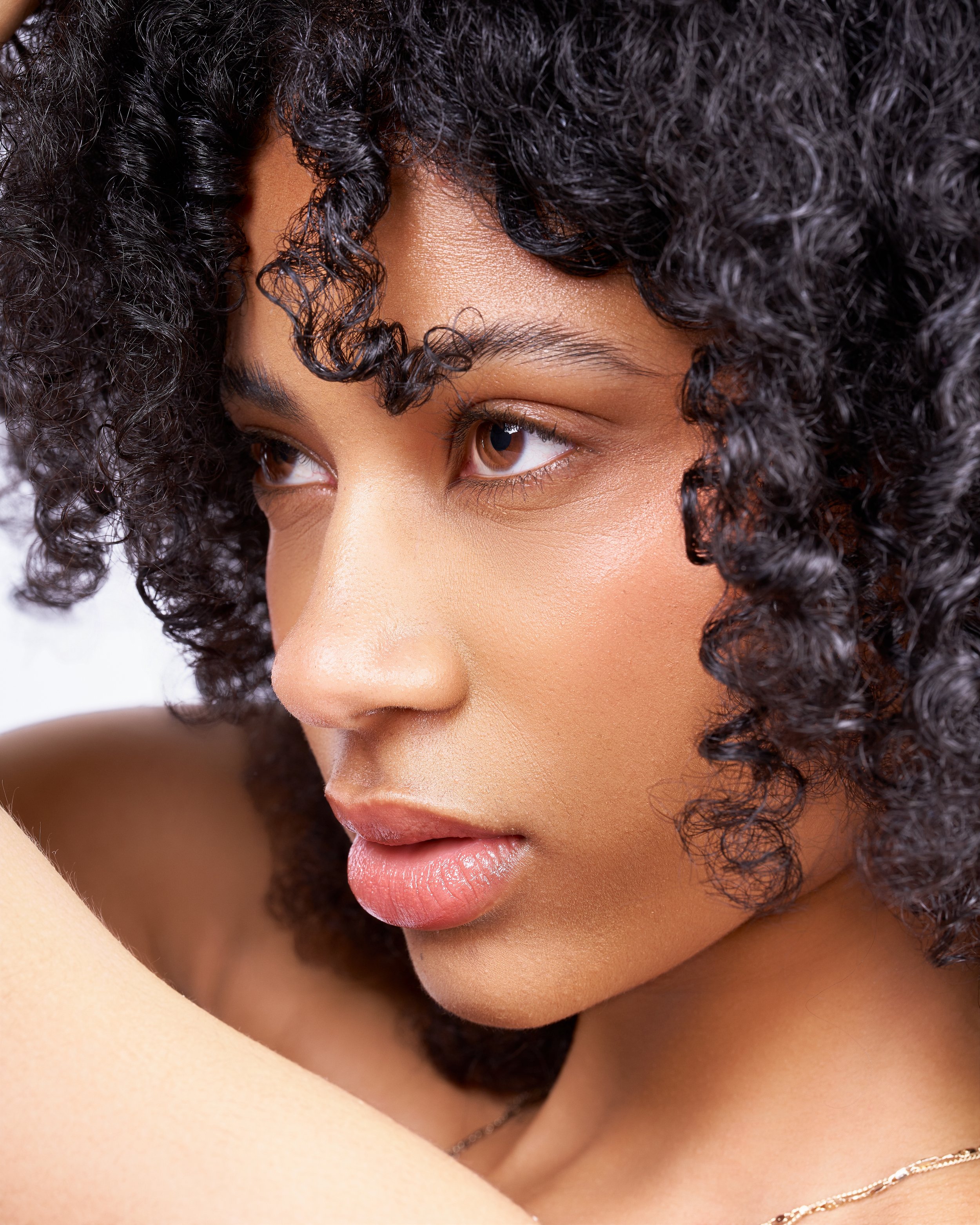 Close-up of a woman with curly black hair, brown eyes, and smooth skin, looking down with a soft expression.