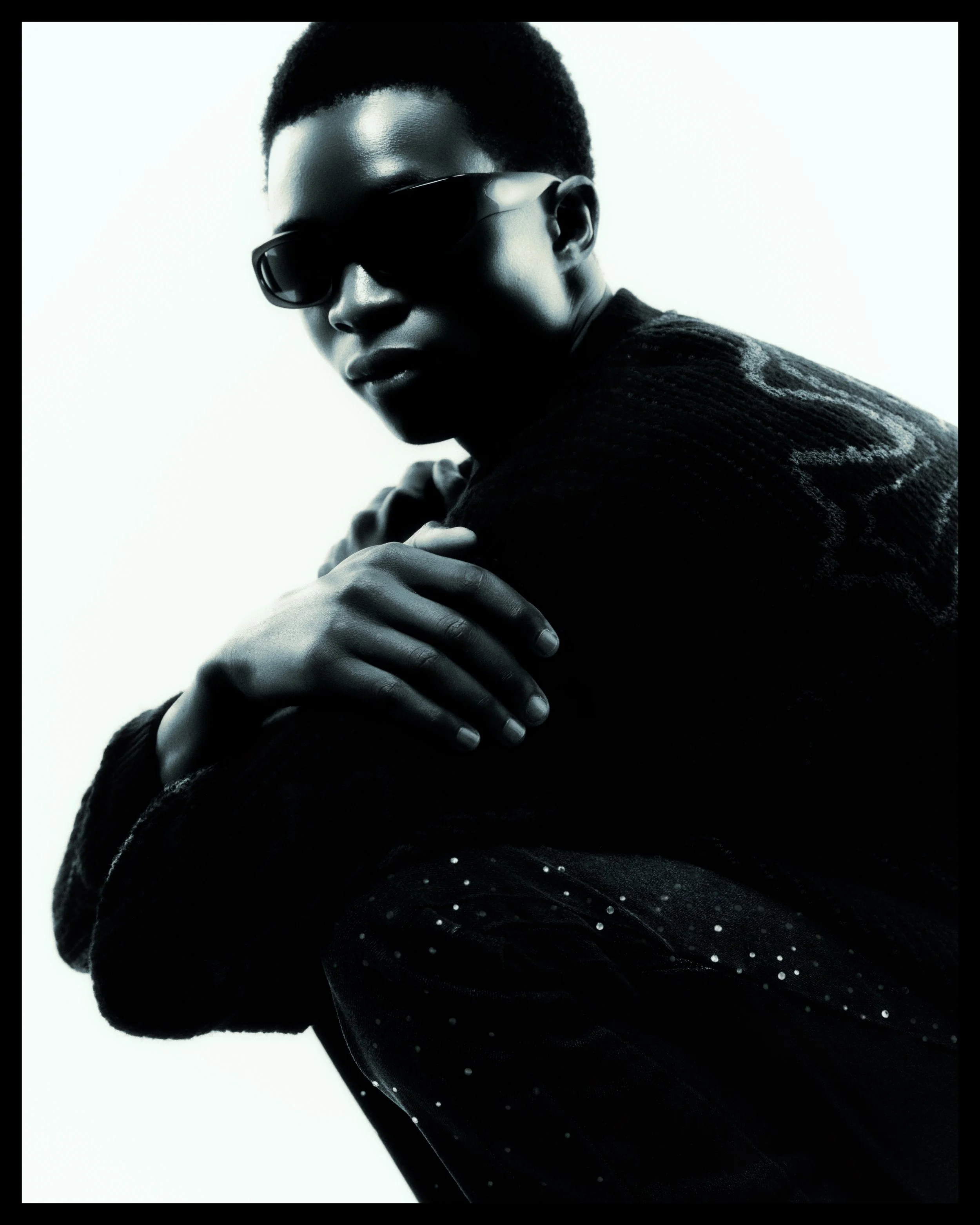 A portrait of a person with short hair wearing dark sunglasses and a dark sweater. The person has their arms crossed and their face turned slightly to the side against a plain light background.