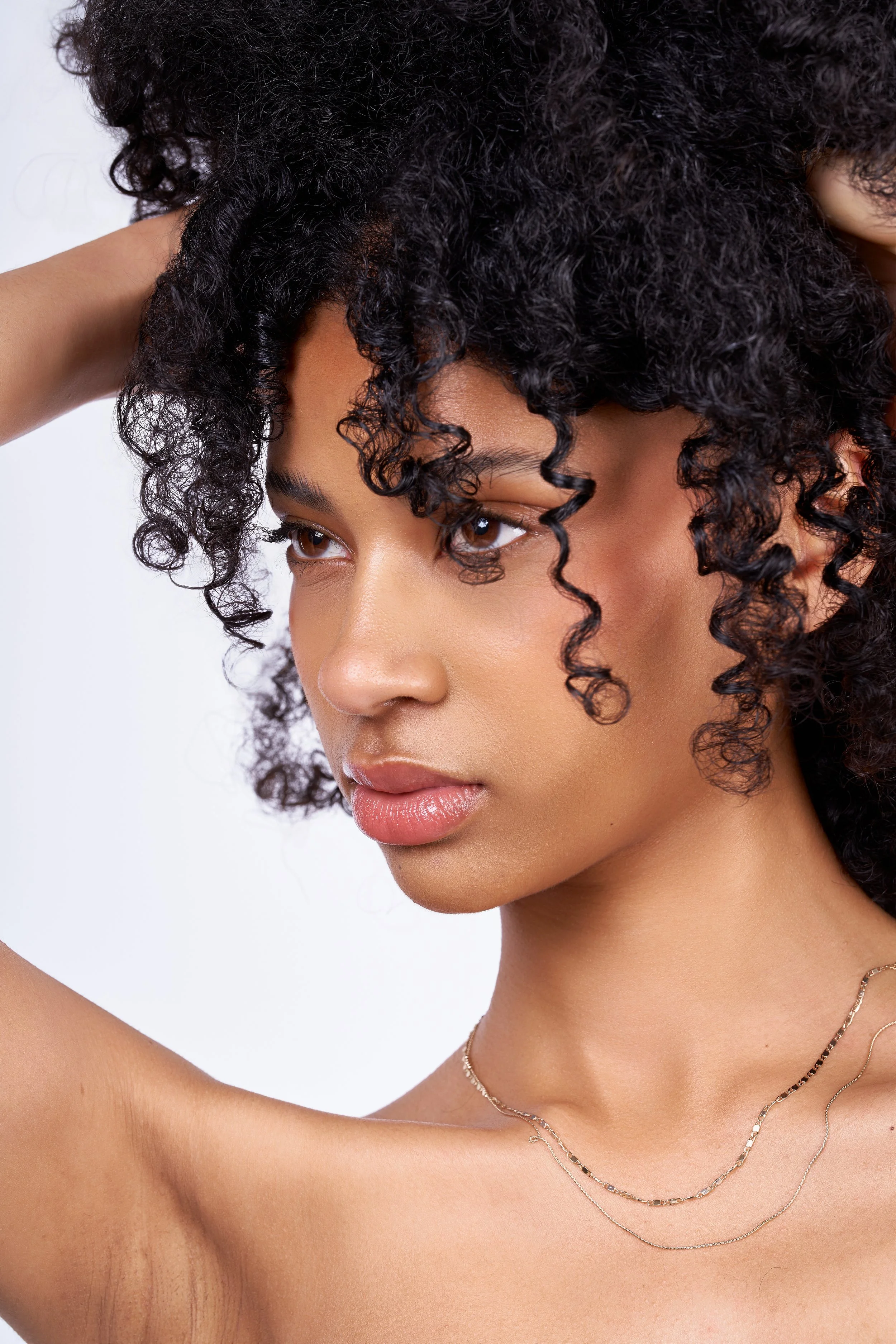 A close-up of a woman with dark curly hair, tan skin, and wearing layered necklaces. She is touching her hair with one hand, and her face is partially visible, showing brown eyes and pink lips.