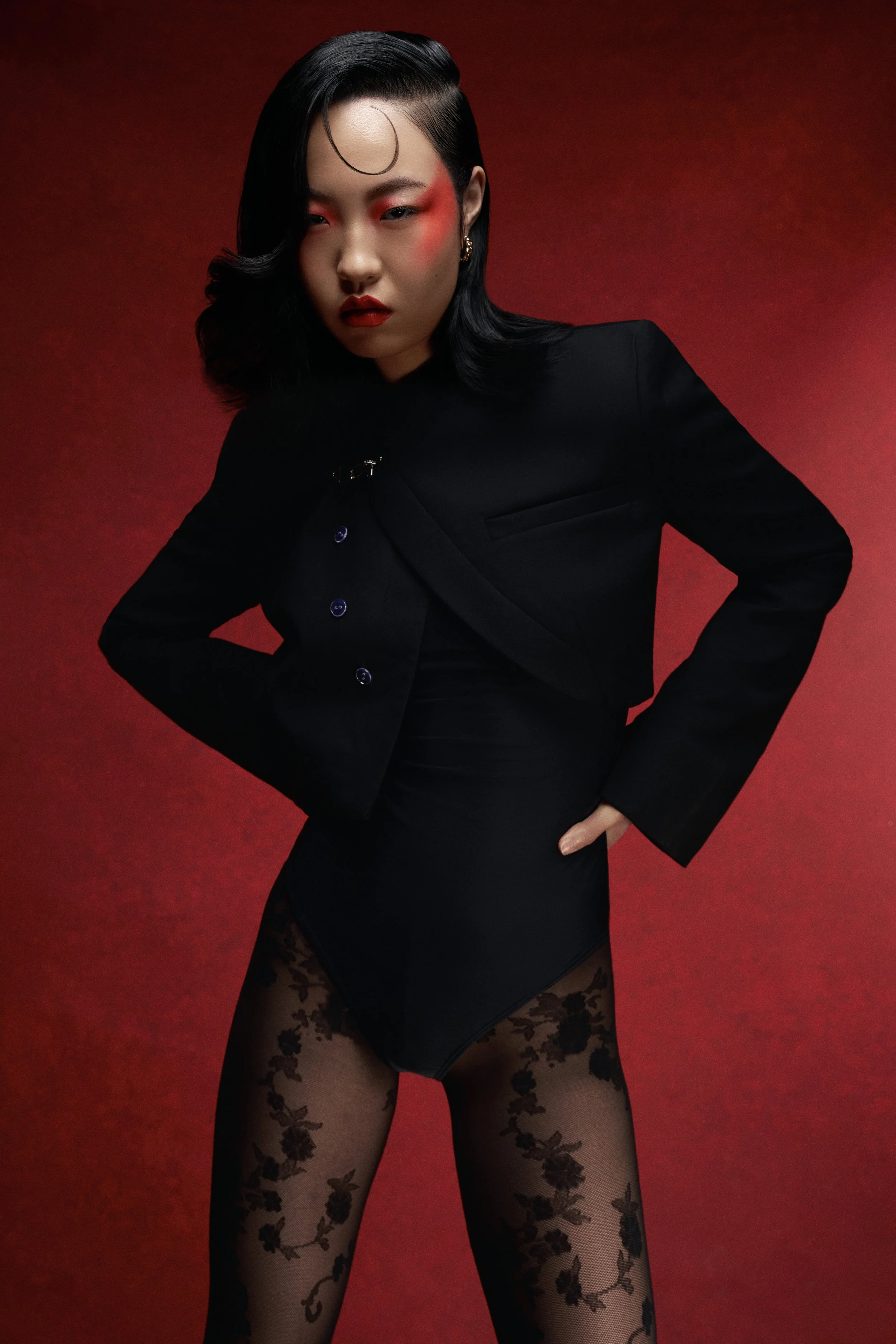 A woman with dark wavy hair styled to one side, wearing a black blazer, black lace stockings with floral patterns, and red makeup, posing against a red background.