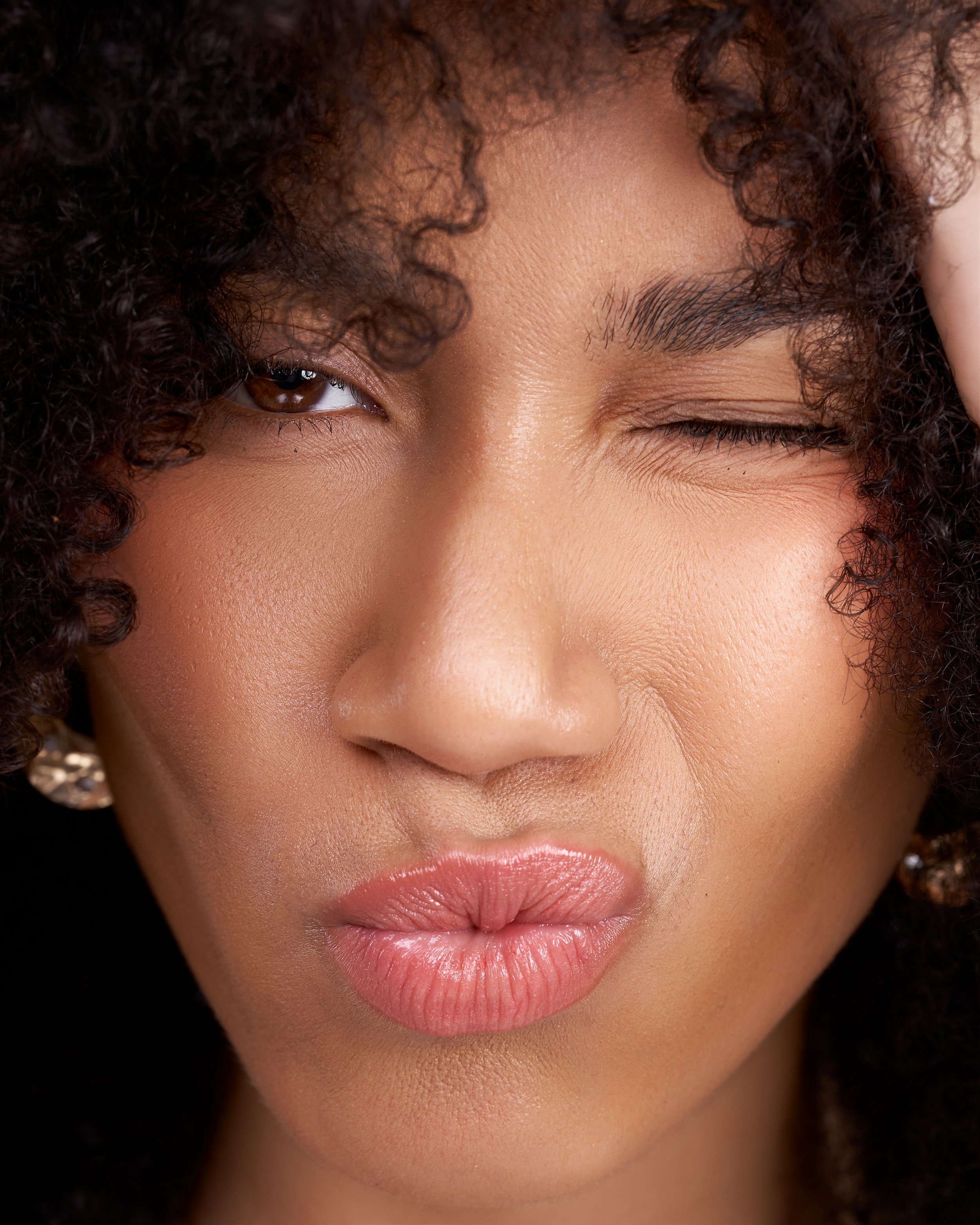 Close-up of a woman's face, with one eye closed and lips pursed, showing curly hair, smooth skin, and well-groomed eyebrows.