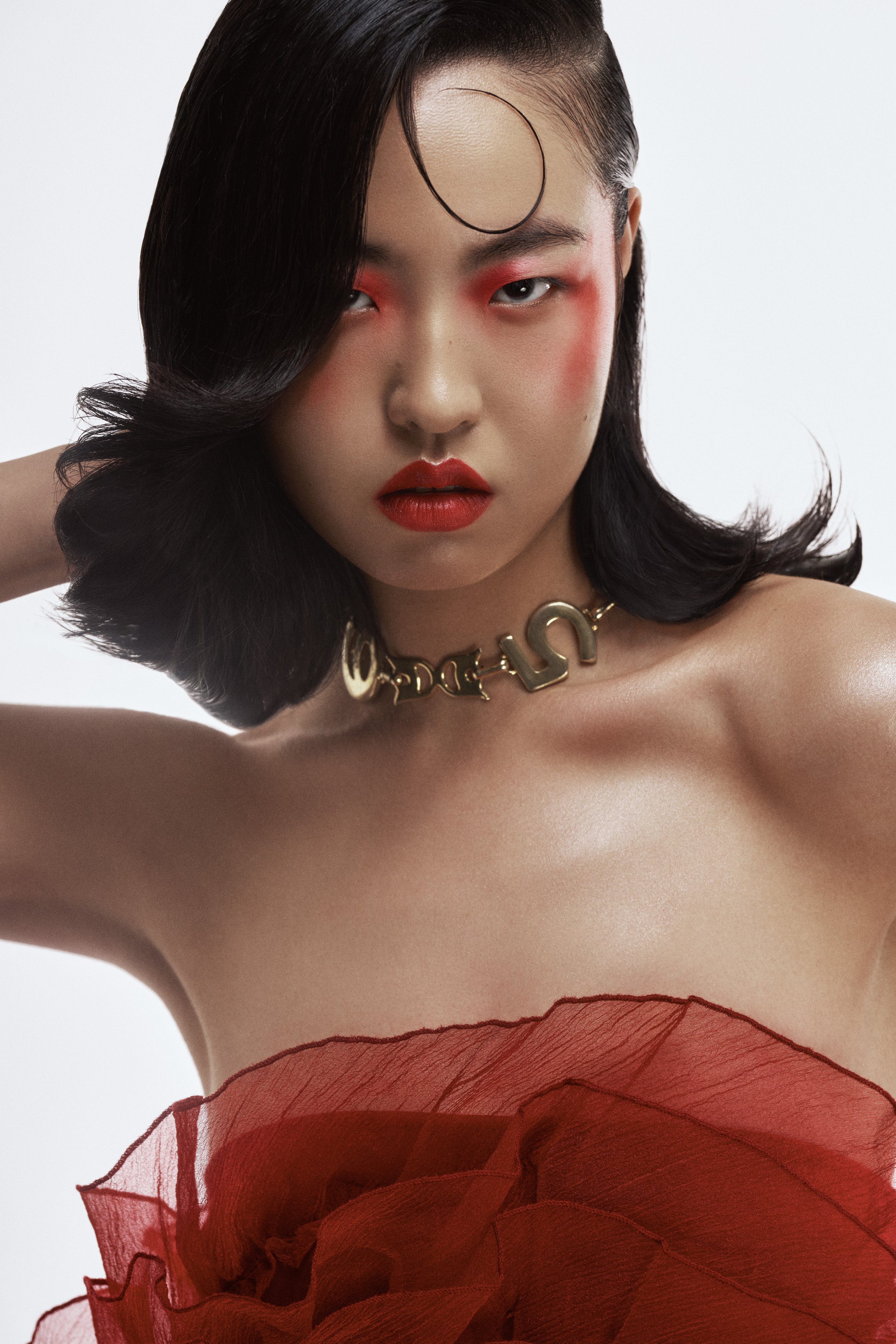 Close-up of a woman with dark hair styled in waves, wearing bold red lipstick and red eye makeup. She is wearing a large gold necklace and a sheer red ruffled fabric draped around her shoulders.