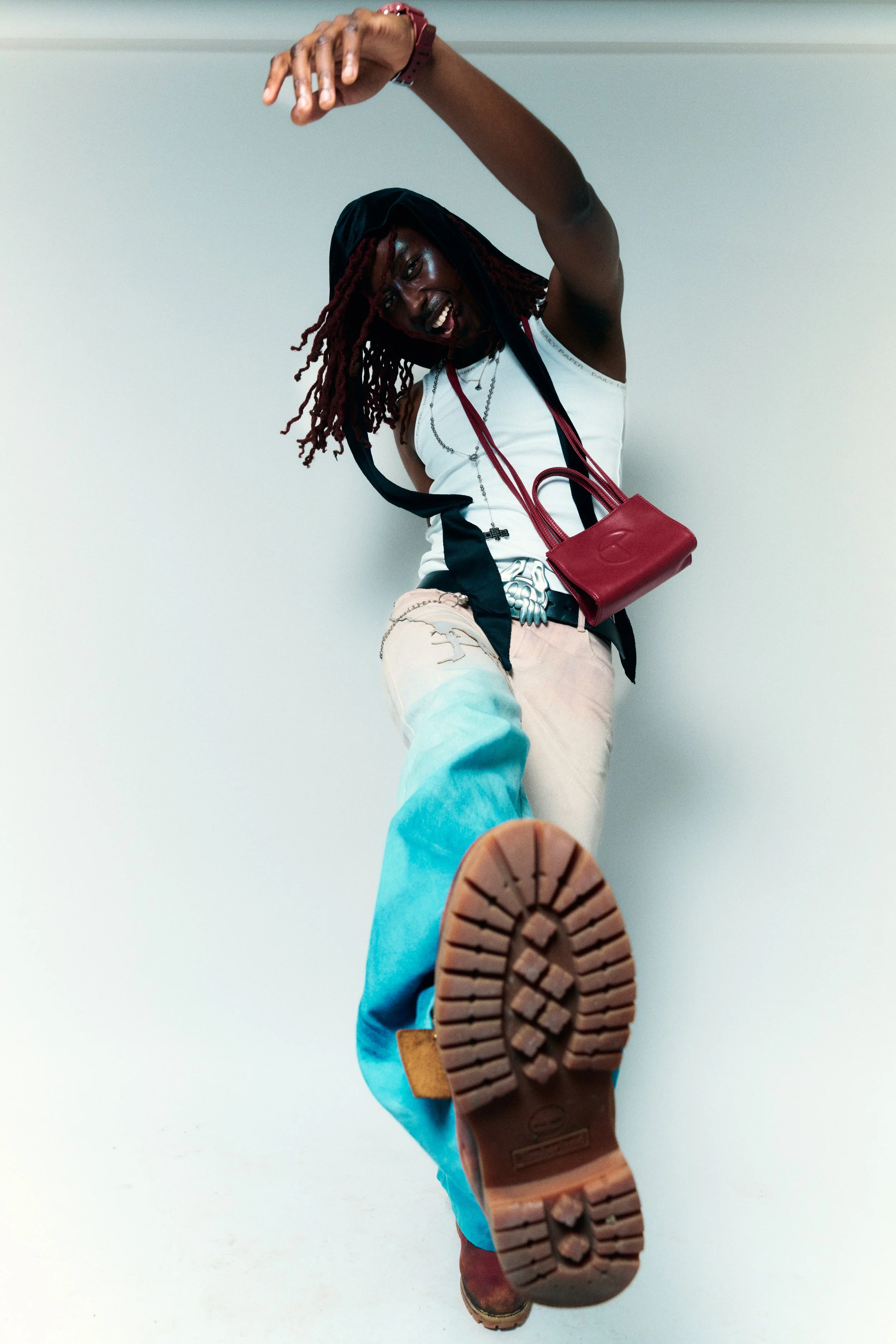 Person standing and kicking towards the camera, wearing colorful denim jeans and a white tank top, with a red purse and accessories, in front of a plain wall.