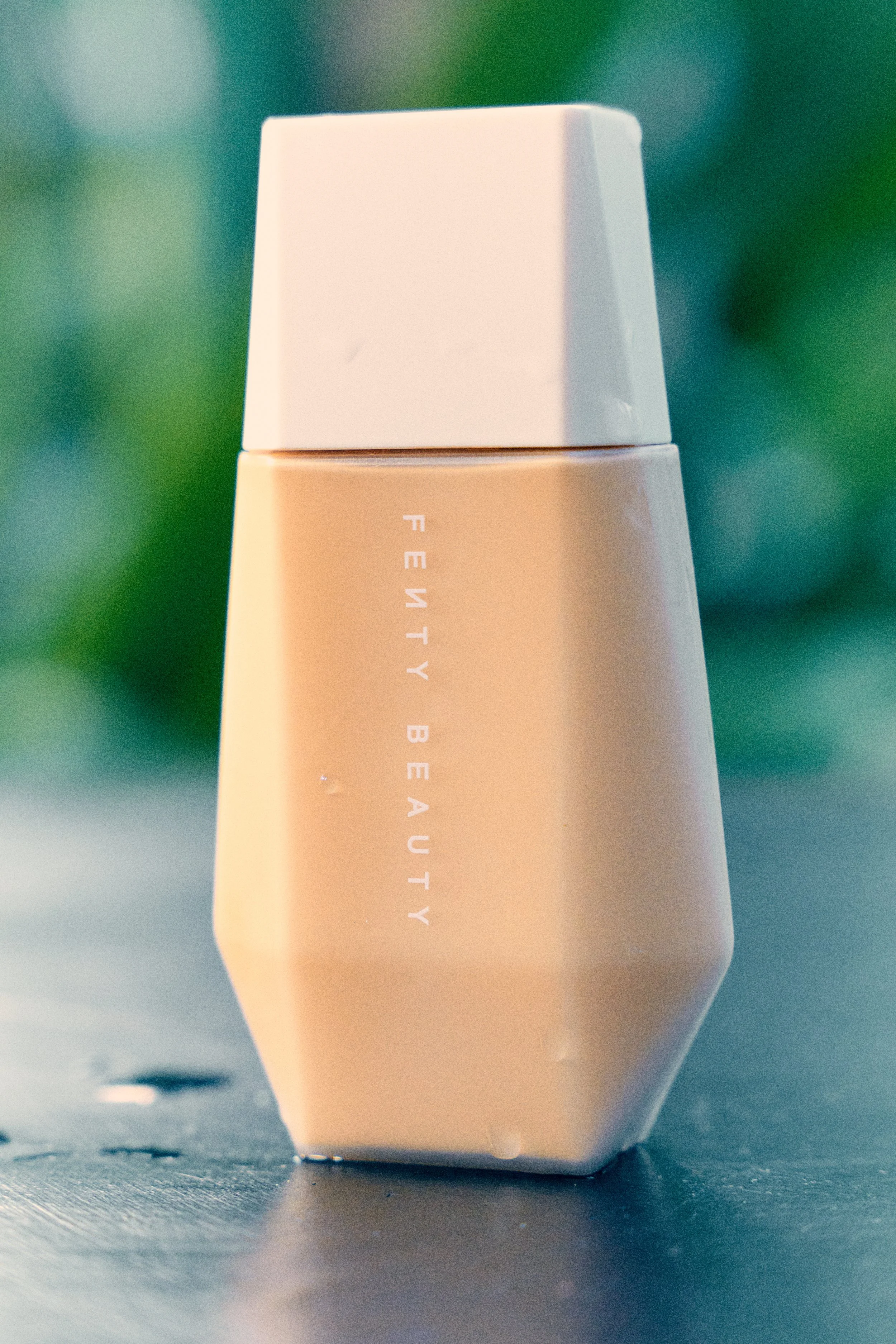 Close-up of a beige bottle with the word 'FEELTY BEA...' partially visible on its side, placed on a dark surface with a blurred green background.