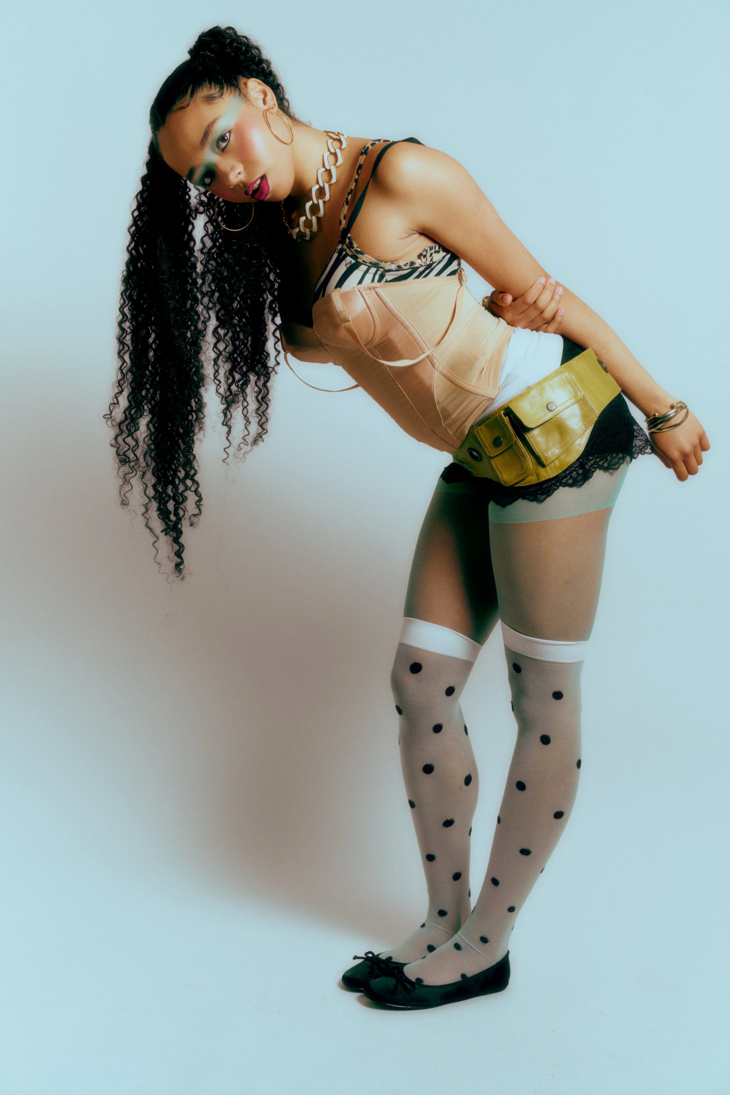 A woman with long curly hair and gold hoop earrings leans to one side against a plain white background. She wears a sleeveless top, a chain necklace, a yellow pouch belt, black shorts with lace trim, polka dot tights, and black shoes.