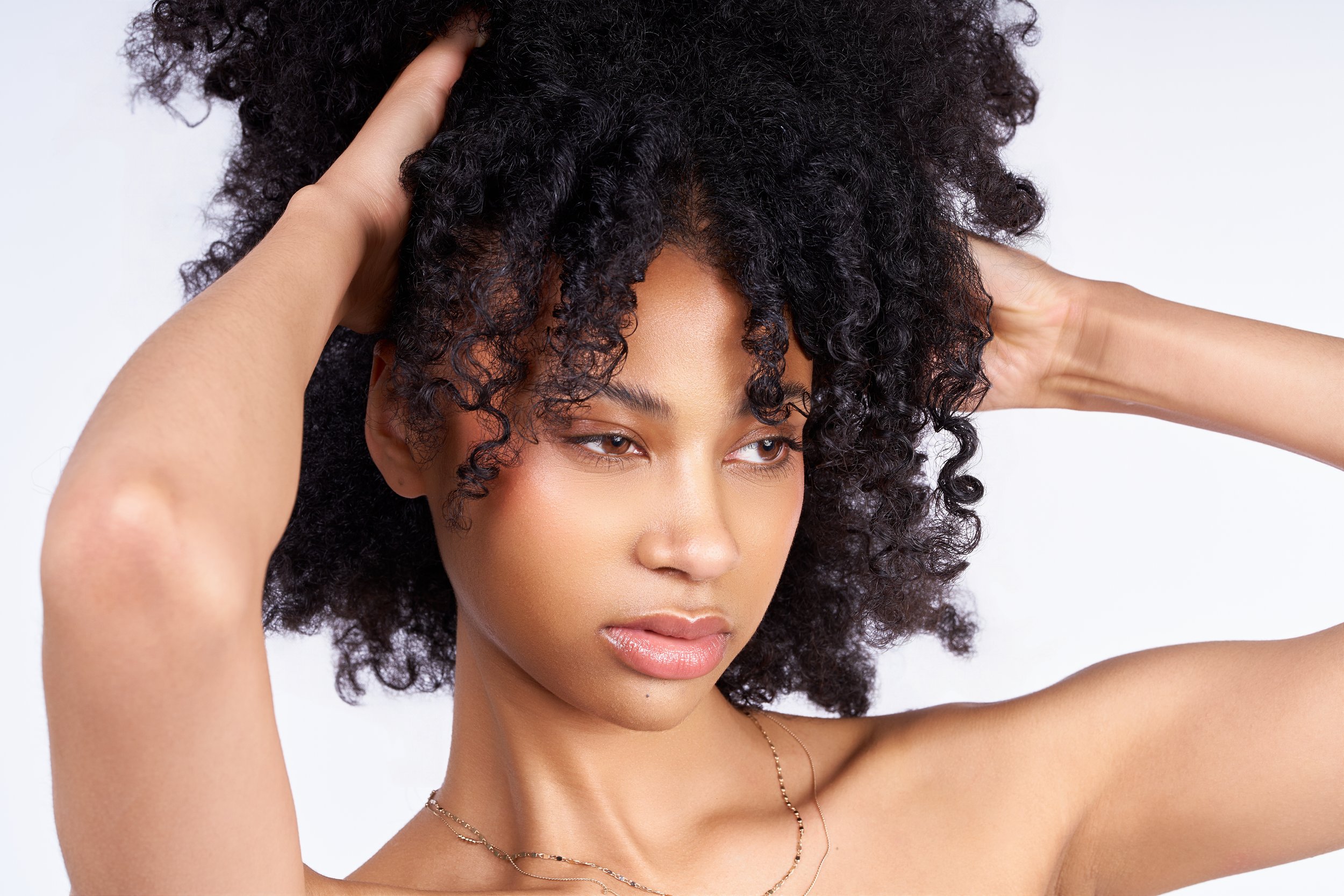 A woman with medium skin tone and natural curly hair, softly holding her hair with both hands, against a plain light background.