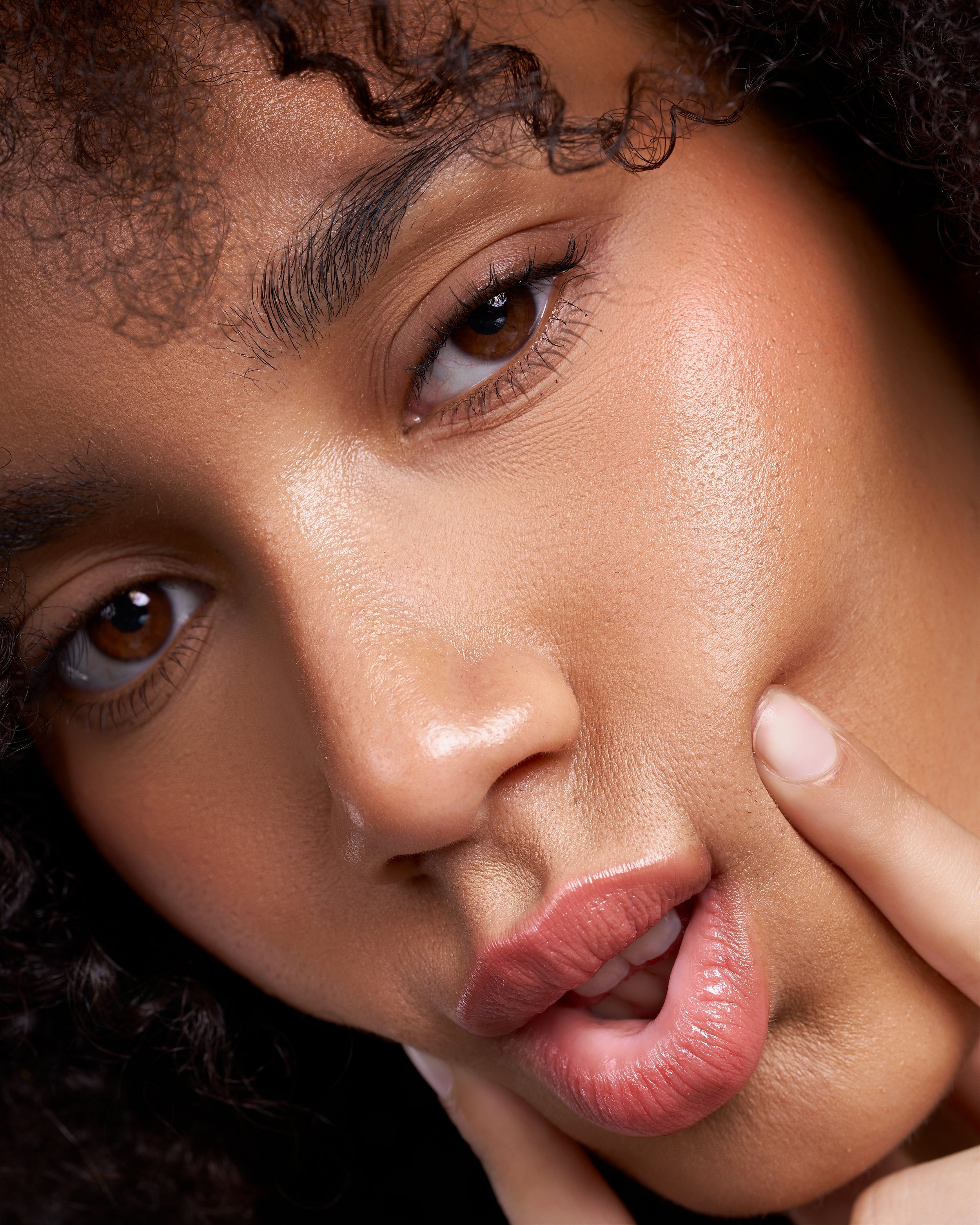 Close-up of a woman with light brown skin, brown eyes, curly dark hair, and full lips, touching her mouth with her finger.