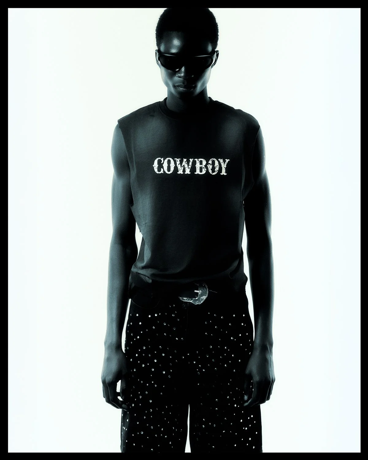 Fashion mannequin wearing a sleeveless black shirt with the word "COWBOY" printed on it, black pants with white polka dots, and dark sunglasses, against a plain light background.
