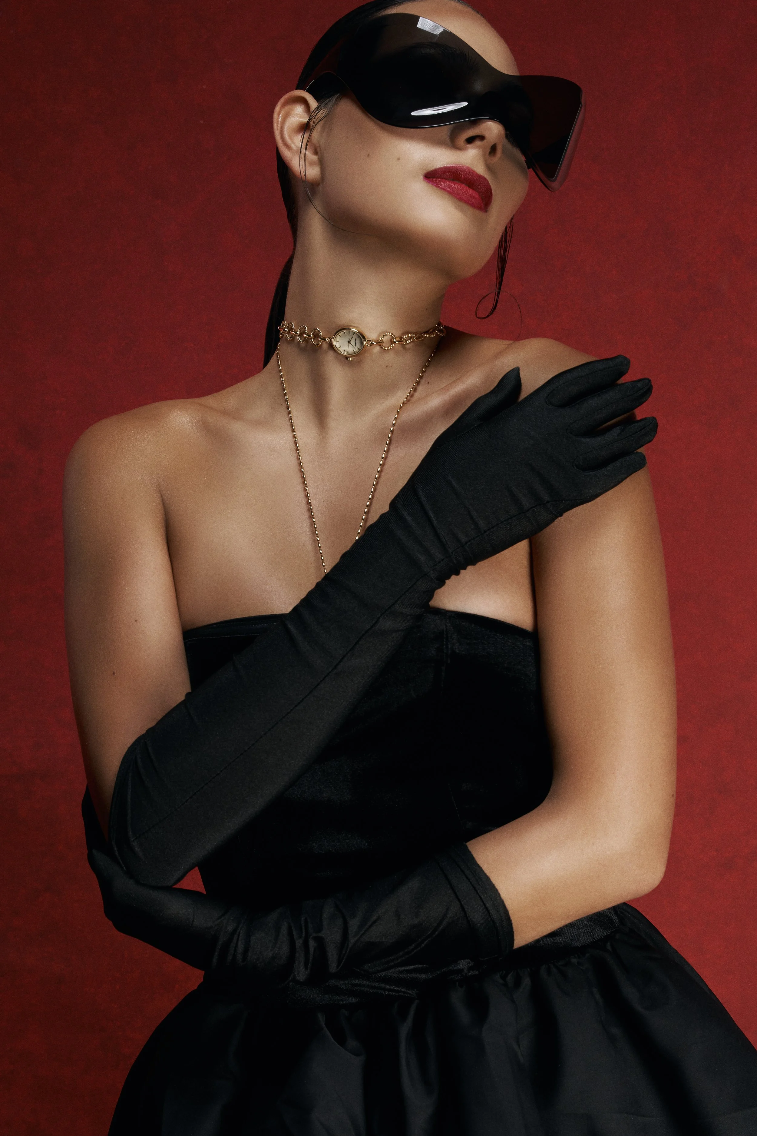 A woman wearing oversized black sunglasses, a gold watch necklace, and black gloves against a red background.