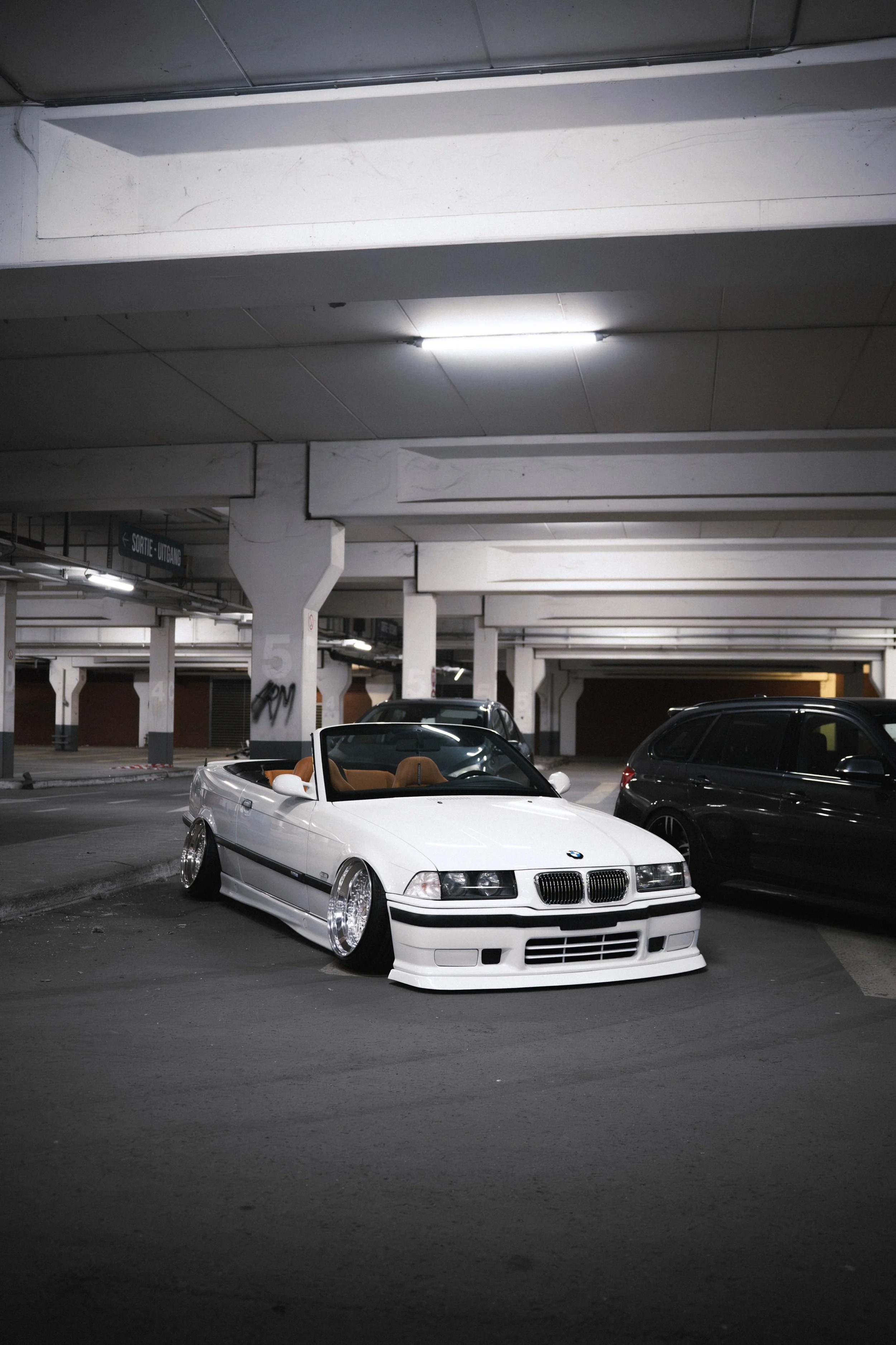 A white BMW convertible with tan interior parked in a dimly lit underground parking garage.