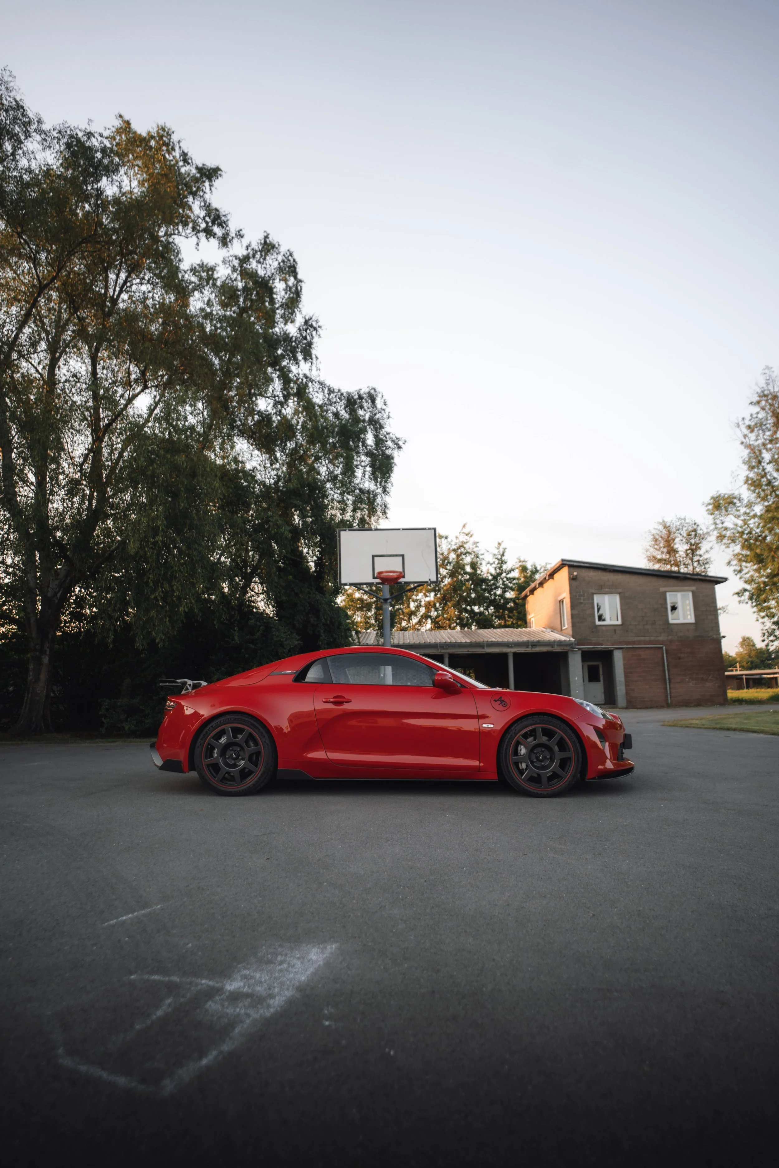 A red sports car parked on a gray asphalt parking lot with a basketball hoop, a building, and trees in the background during evening.