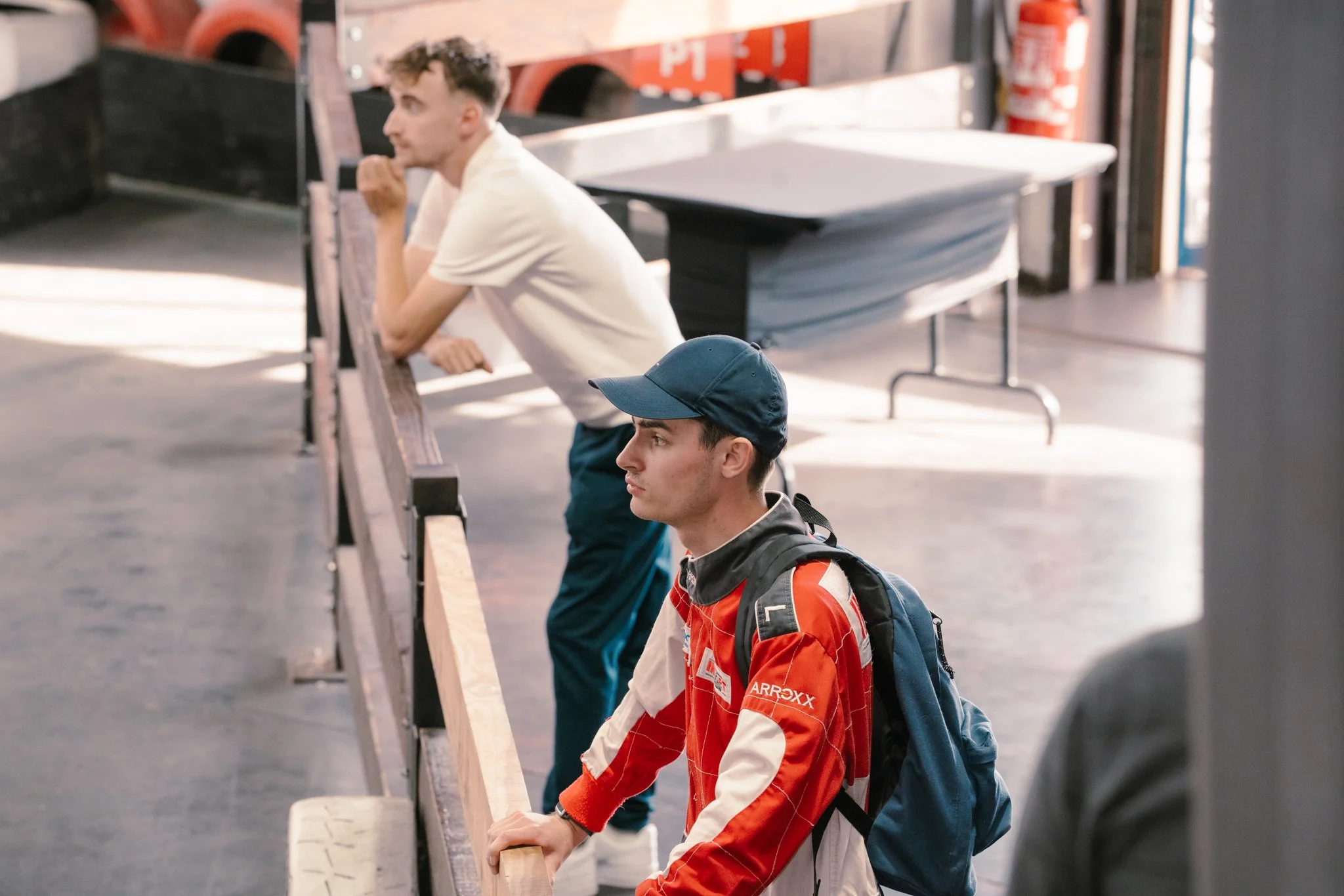 Two young men standing at a barrier, looking towards the left. One is wearing a white shirt, and the other is wearing a red and white jacket with a dark backpack, a navy blue cap, and leaning on the barrier.