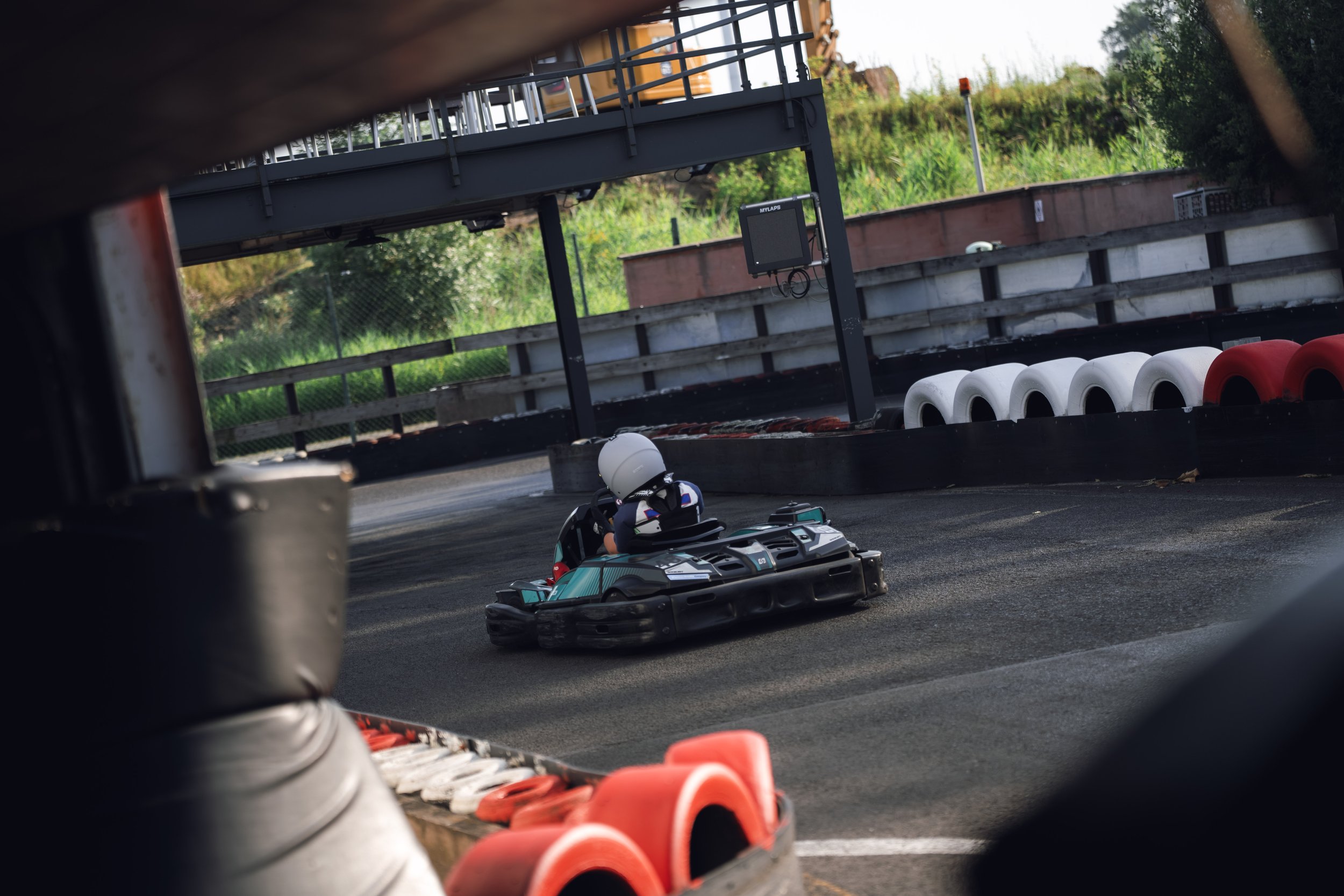 Kids riding go-karts on a race track with safety barriers, seen through the frame of a tire.