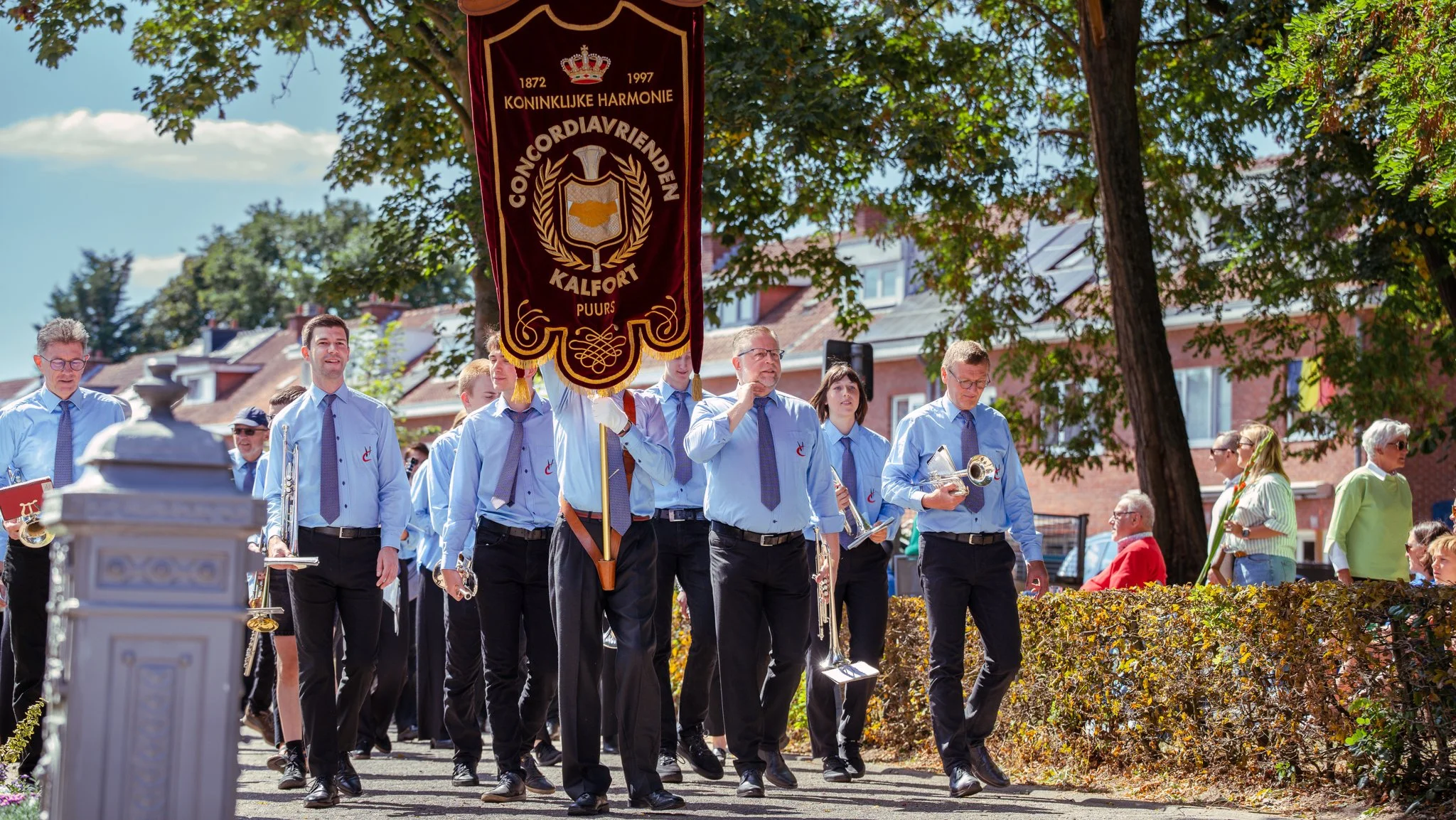 A marching band dressed in light blue shirts and dark pants parades outdoors with some members playing instruments such as trumpet and clarinet, while one member holds a large decorative banner with crest and text. People watch the parade from the si