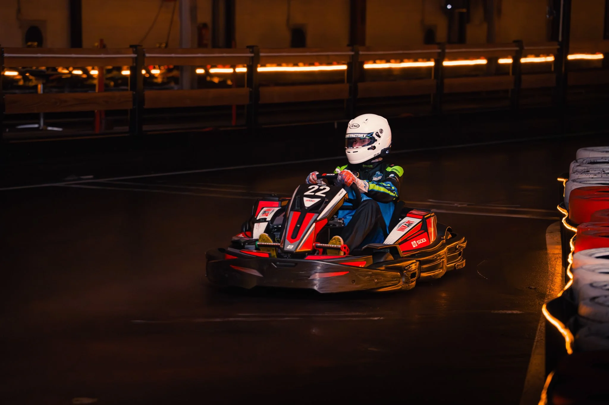 A person driving a go-kart on an indoor racing track, wearing a white helmet and a racing suit, illuminated by warm lighting.