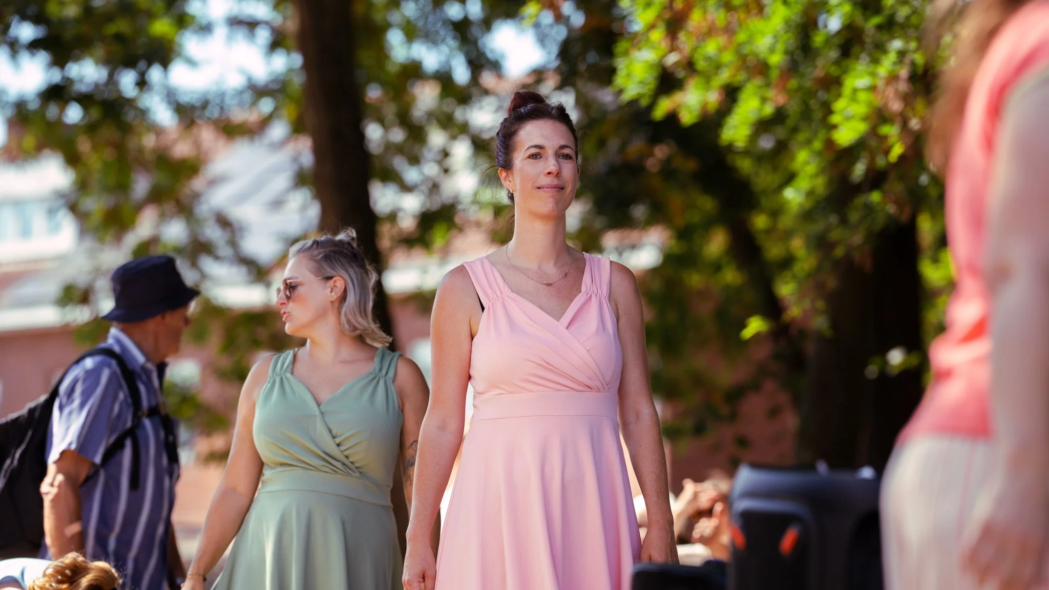 Women and a man standing outdoors with trees in the background, some women are dressed in pastel-colored dresses, and a man is wearing a striped shirt and a hat.