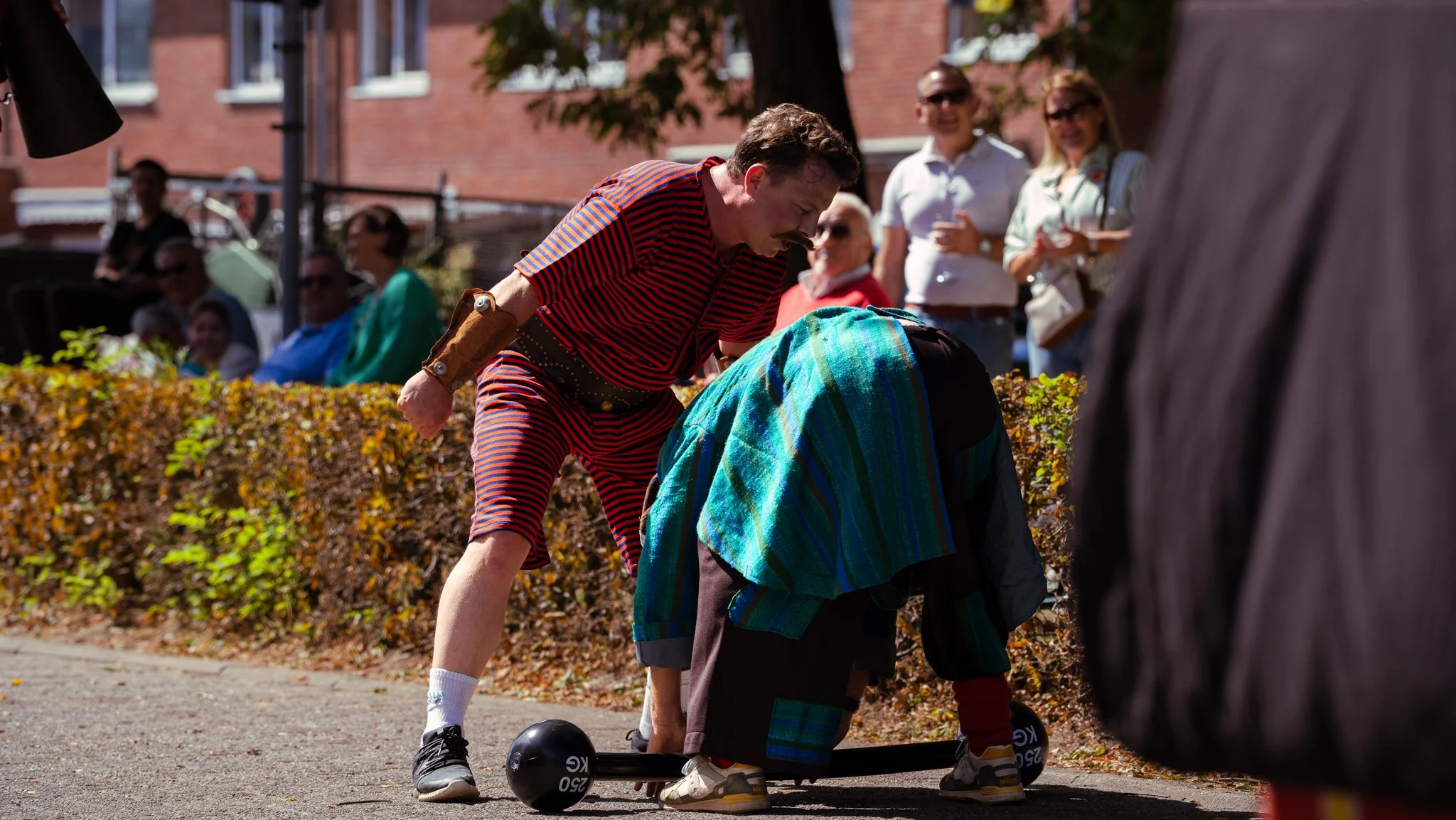 A man dressed in a colorful striped outfit assisting a person in a colorful patchwork costume on a skateboard at an outdoor event, with spectators in the background.