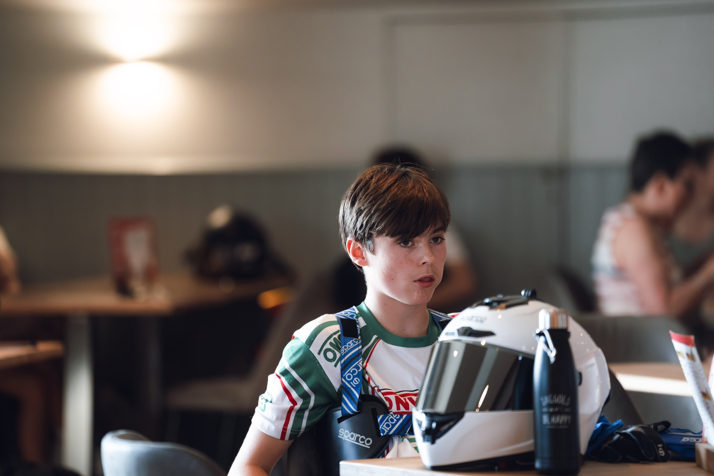 A young boy with brown hair and light skin wearing a green and white racing shirt, sitting at a table with a racing helmet and a simulated racing wheel in front of him, in a room with other children in the background.