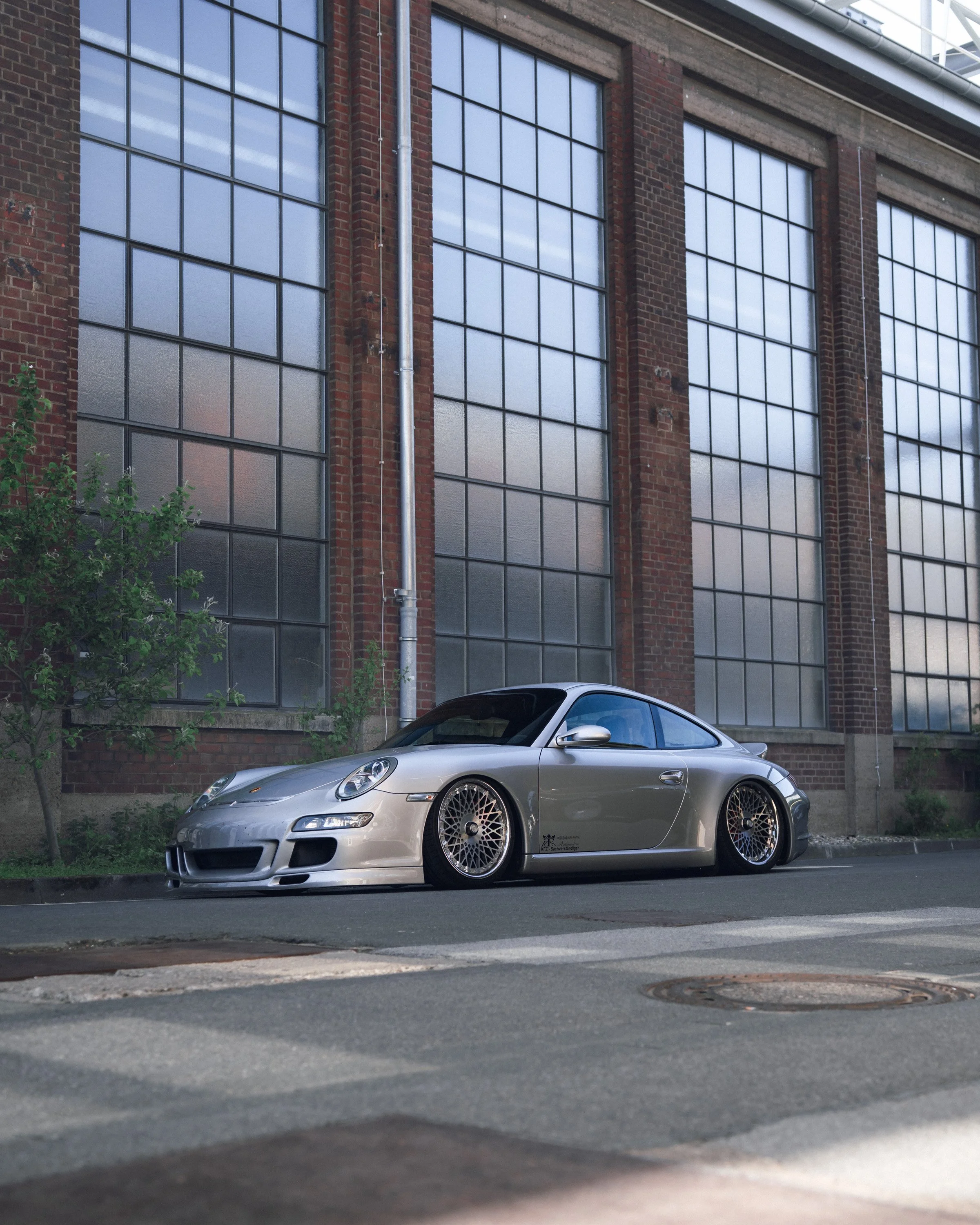 A silver sports car is parked on the street in front of a brick building with large windows and metal pipes. Trees and sidewalk are visible in the scene.