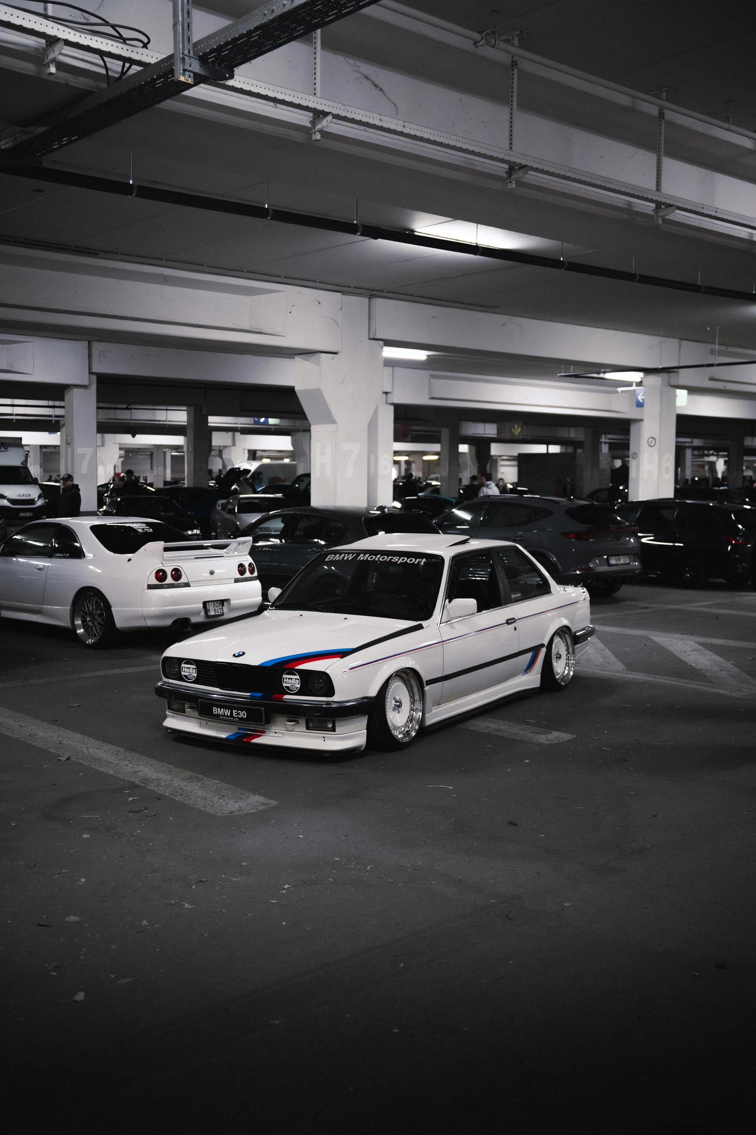 A white BMW E30 with BMW Motorsport livery parked in an underground parking garage, surrounded by other vehicles in dim lighting.