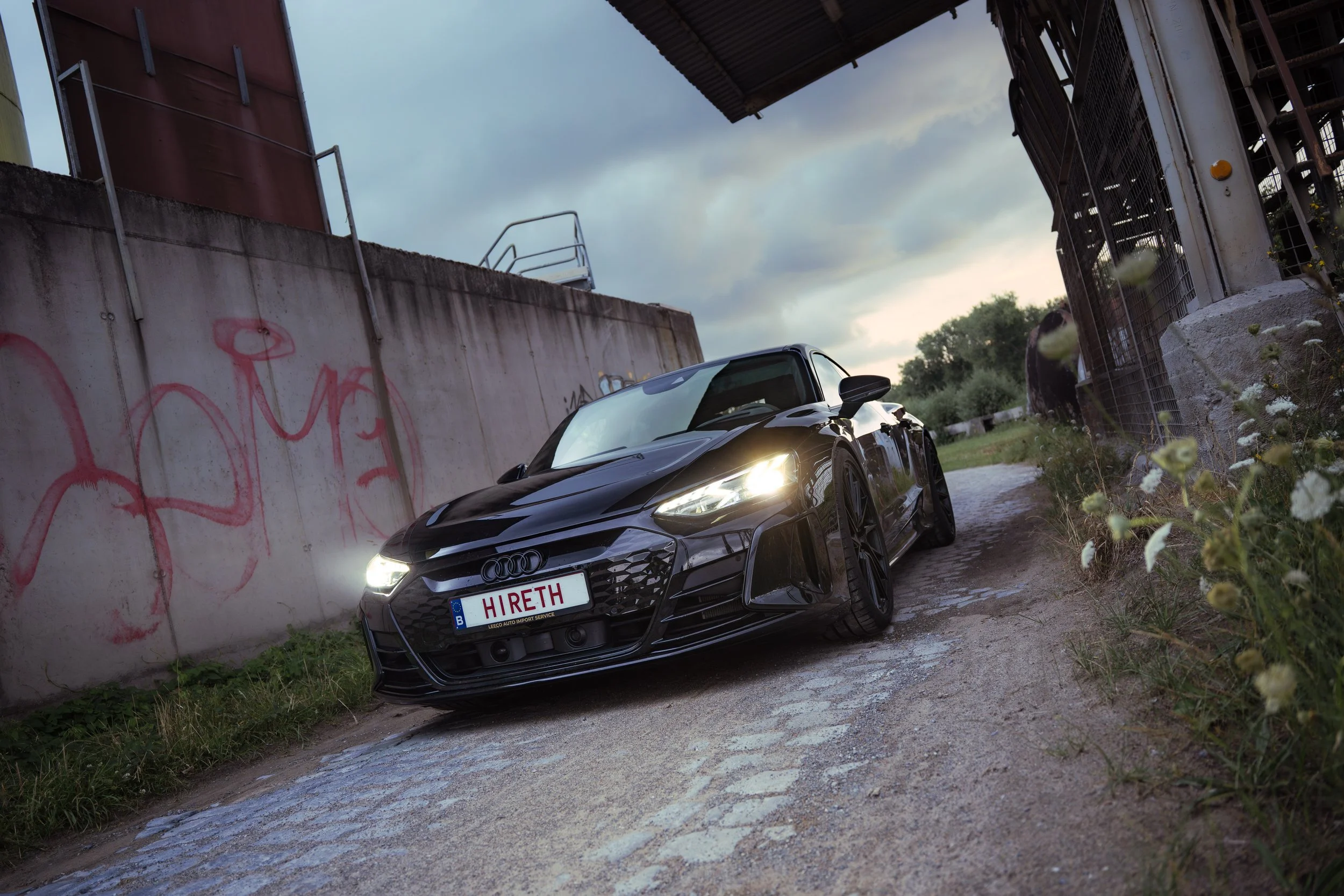 A black Audi sports car parked on a gravel path under a bridge, with graffiti on a concrete wall on the left and an open field with cows in the background.