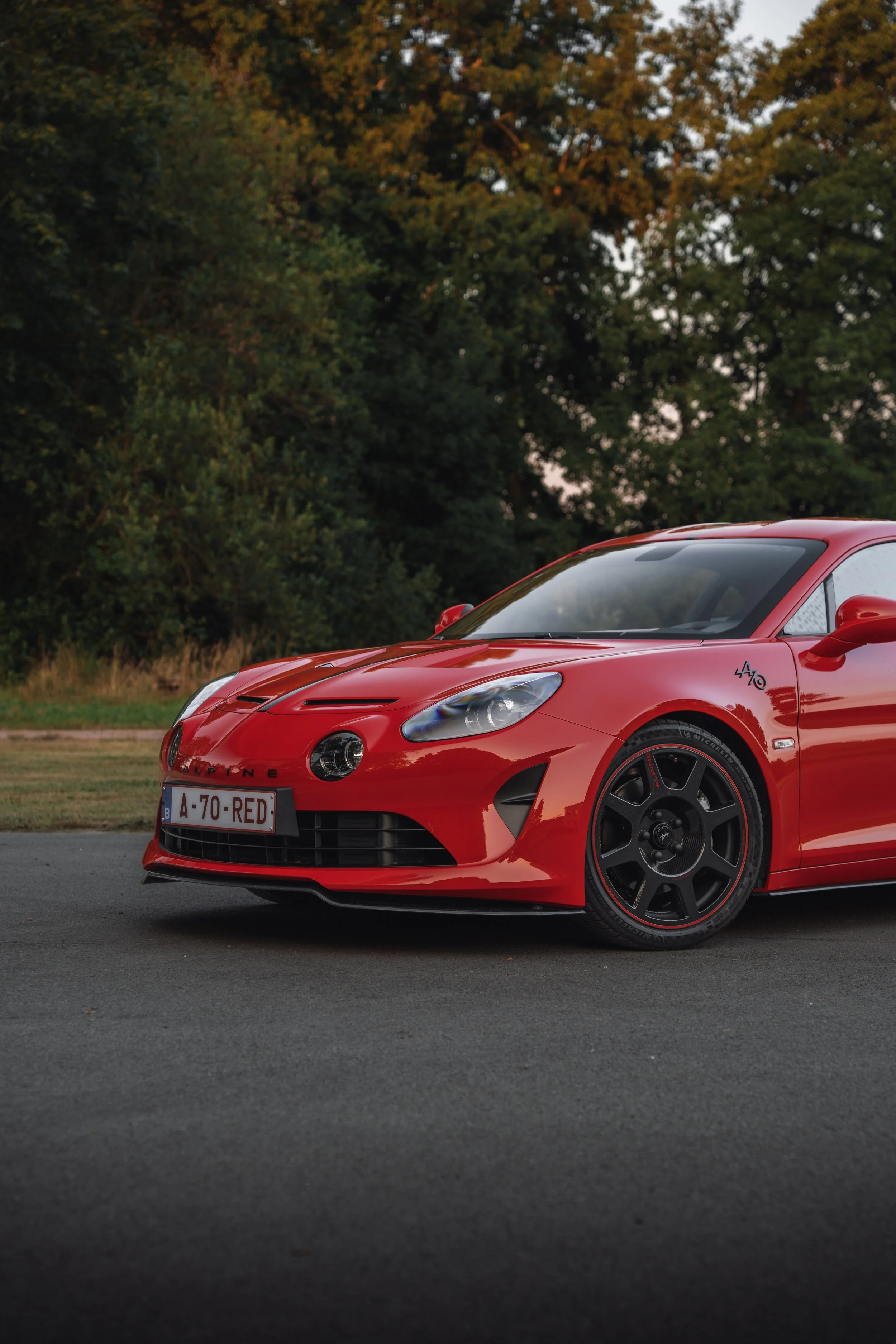 Red sports car parked on a paved surface with a background of green trees and a sunset sky.
