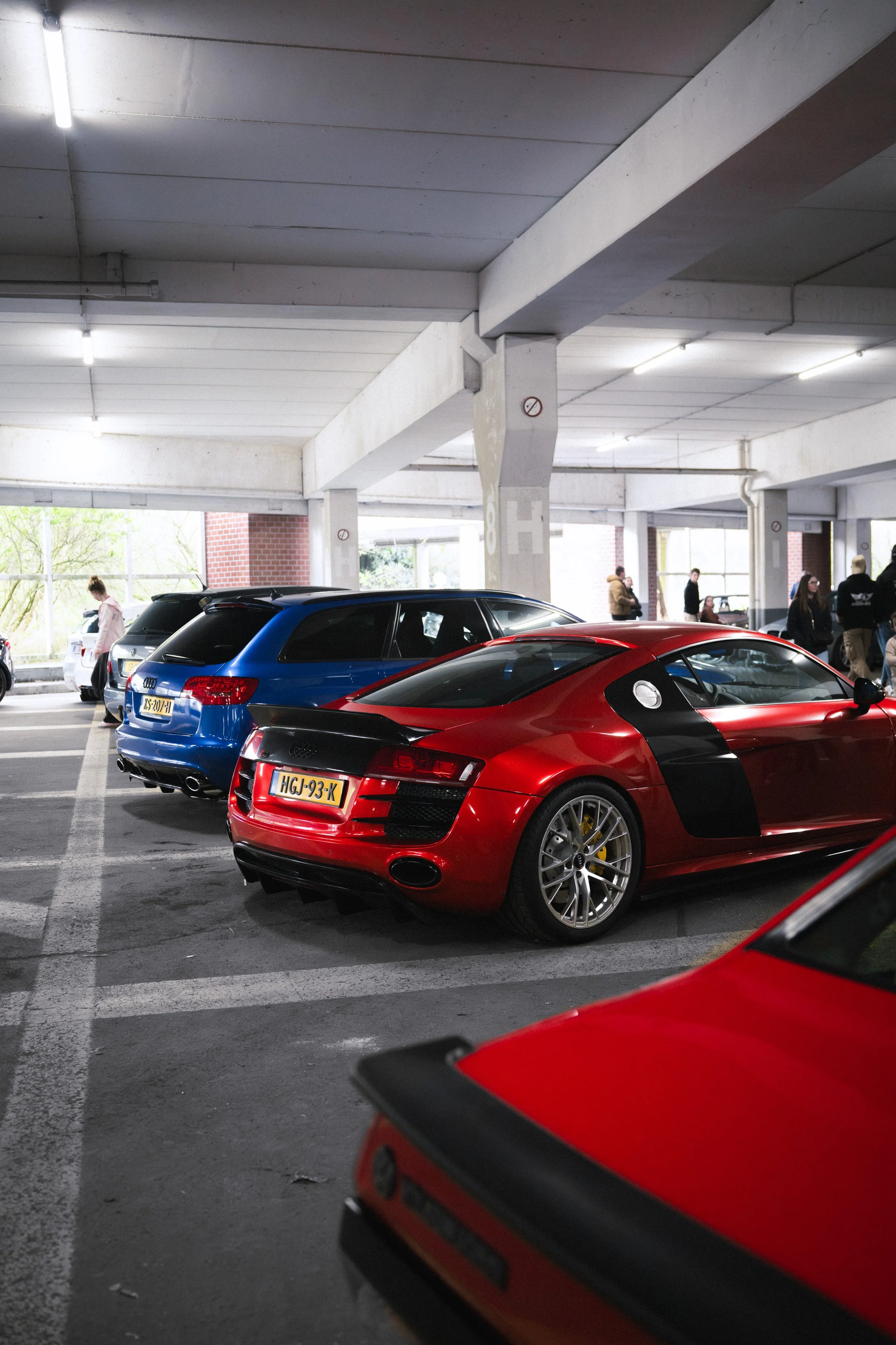 Red sports car parked in a multilevel parking garage with people walking in the background.