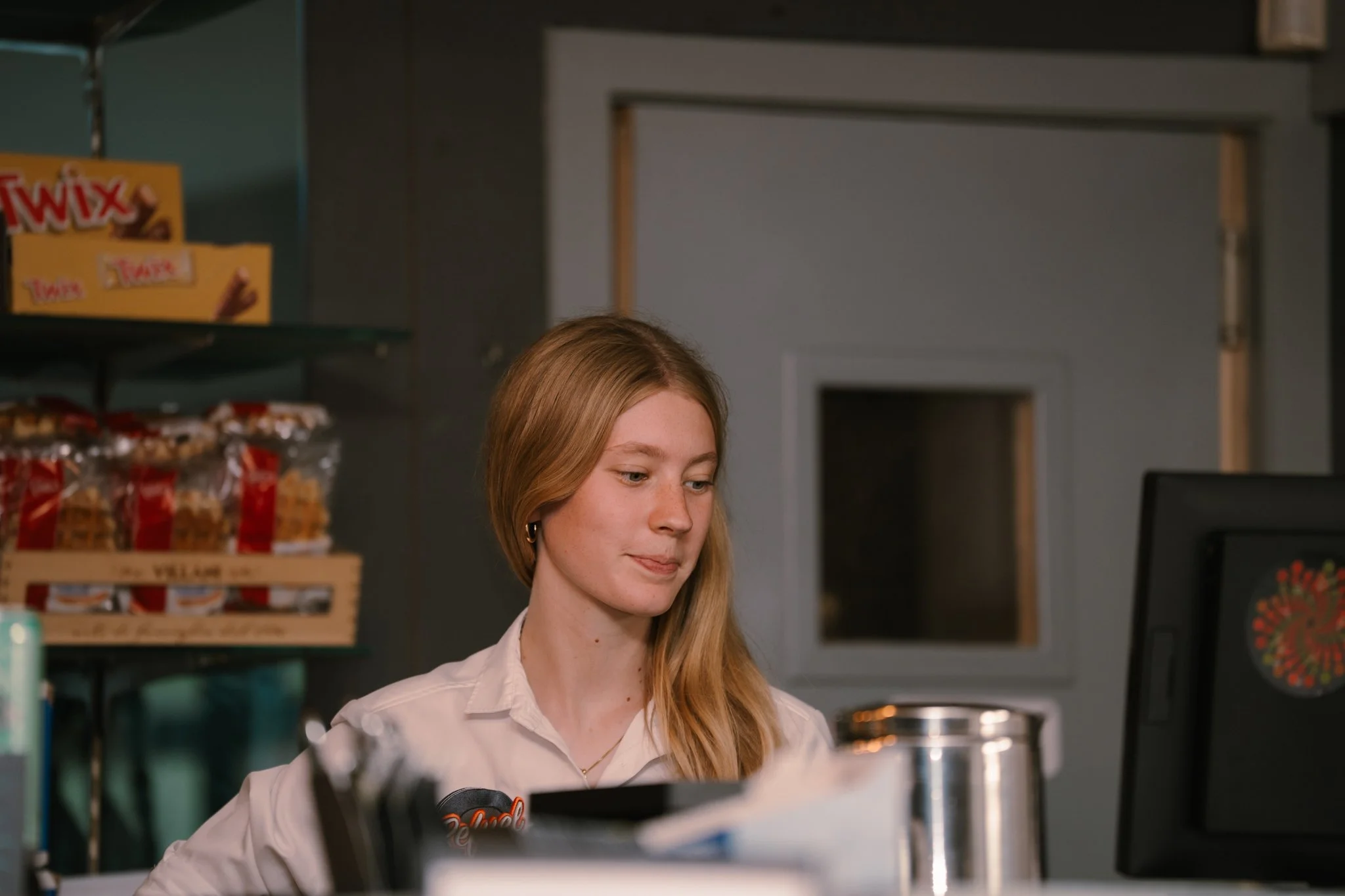 A young woman with long blonde hair, wearing a white uniform, sitting at a desk in what appears to be a retail or convenience store.